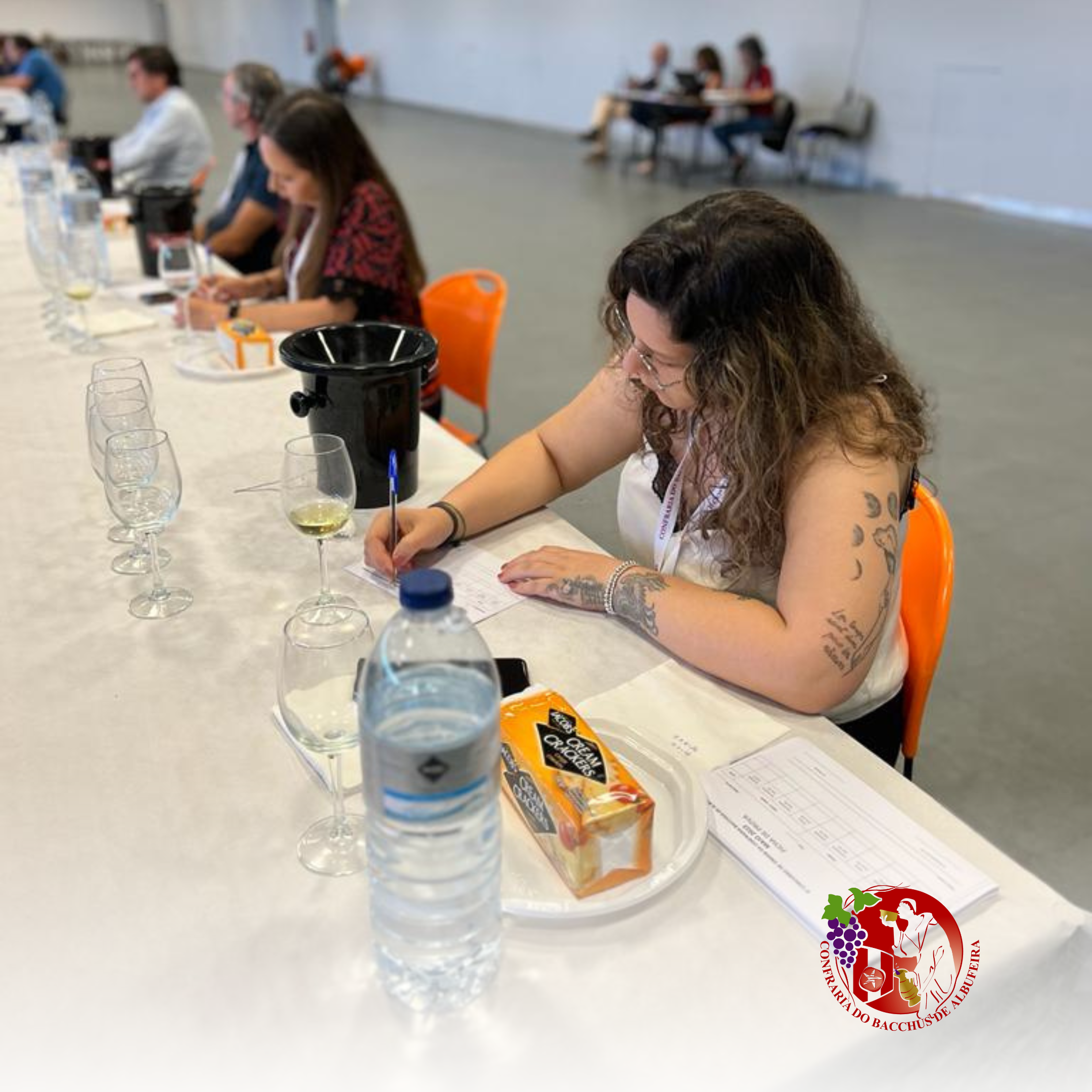 Mulher com tatuagens escrevendo em uma folha na mesa, participando de uma degustação de vinhos, ao lado de garrafas de água, copos de vinho, uma caixa de queijos e papéis de avaliação, com outras pessoas ao fundo em ambiente de conferência.