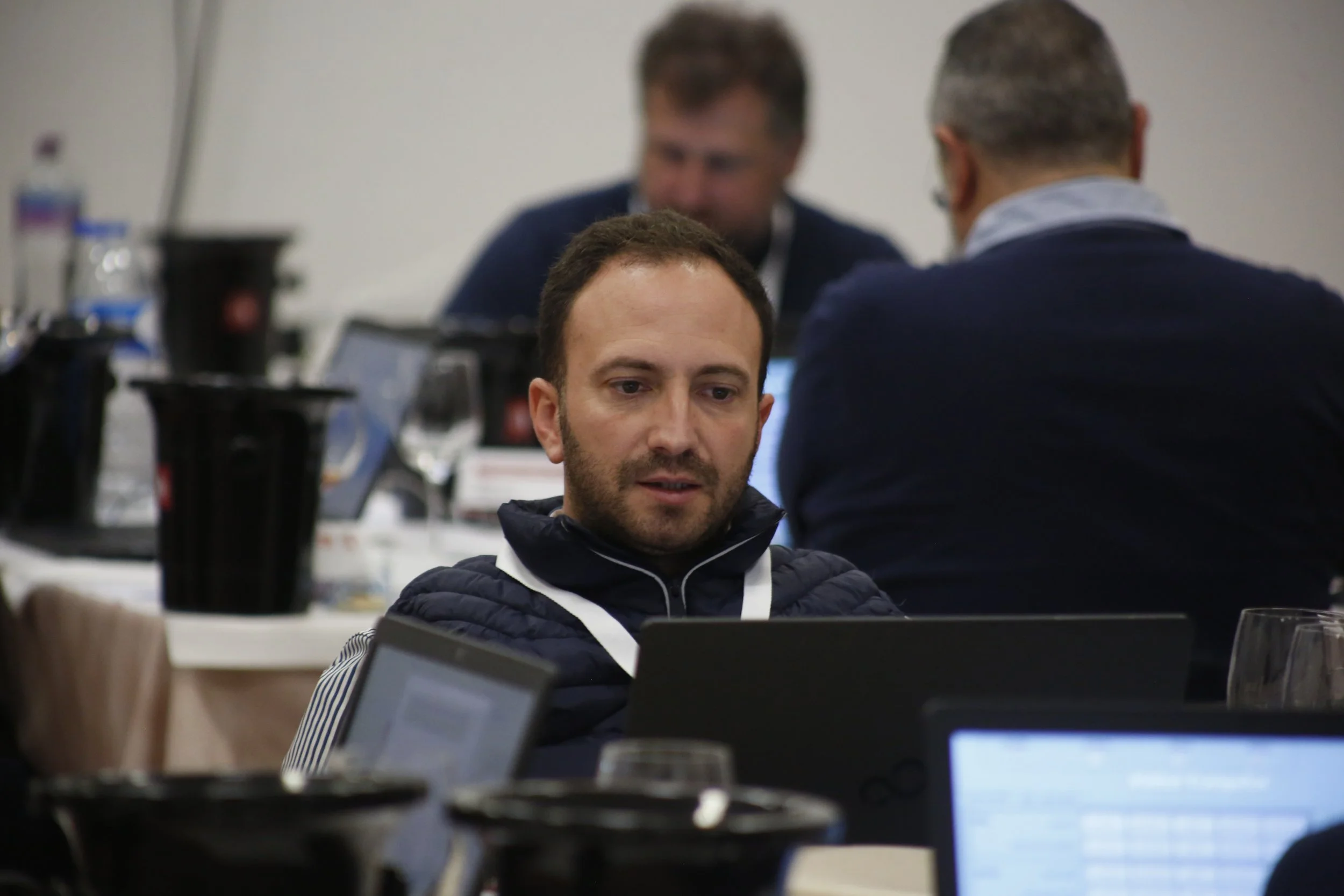 Homem jovem com barba, usando jaqueta preta, sentado em uma mesa de conferência, olhando para o computador, com outras pessoas ao fundo.