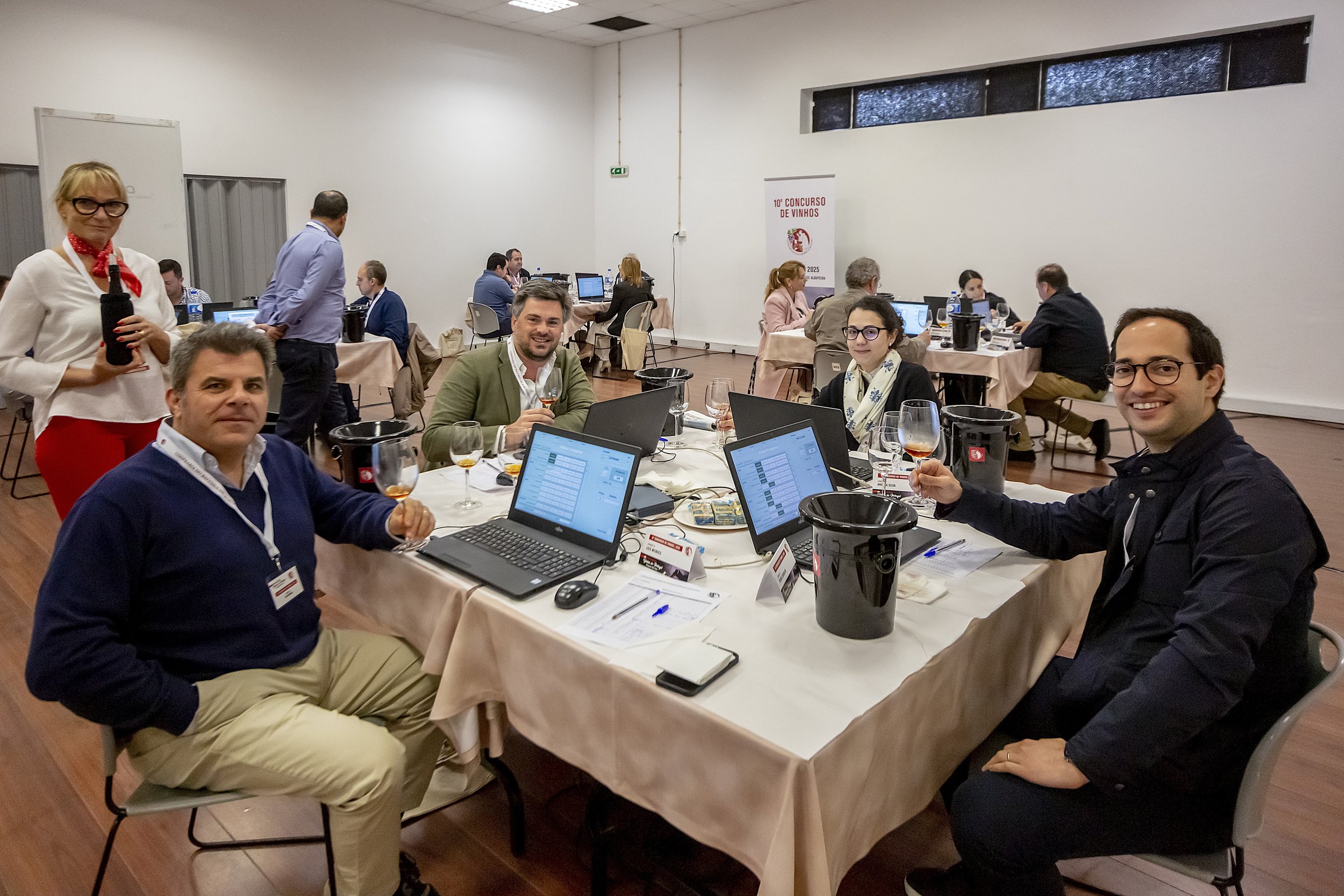 Pessoas participando de um concurso de vinhos em uma sala de conferências com computadores, taças de vinho e uma placa de sinalização no fundo que diz '10º Concurso de Vinhos'.