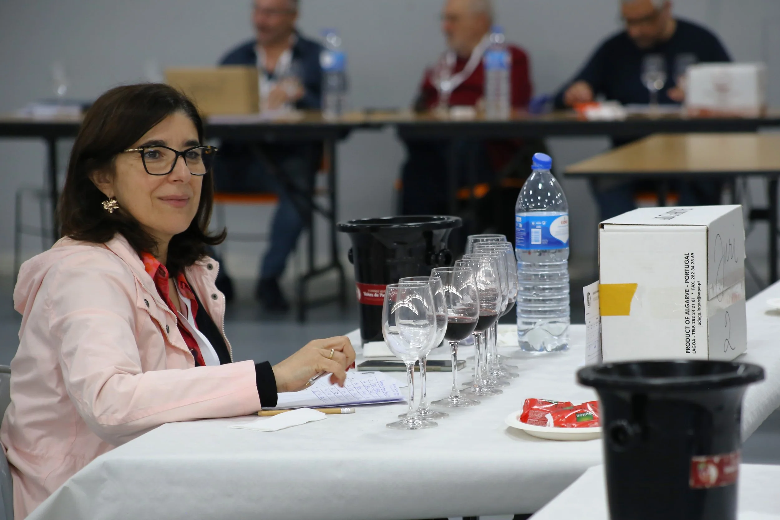 Mulher com óculos sentado em uma mesa com taças de vinho tinto, uma garrafa de água e uma caixa, em ambiente de reunião ou evento.