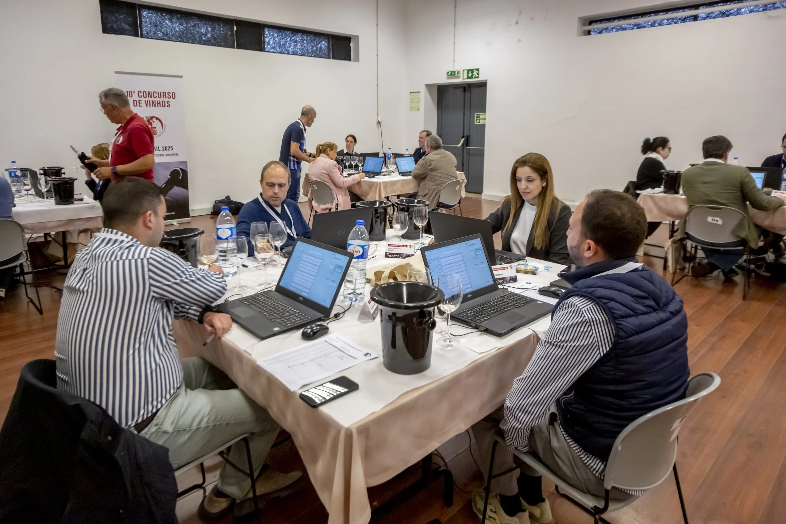 Participantes em uma reunião de concurso de vinhos, sentados ao redor de uma mesa com laptops, garrafas de água e copos, em um salão com paredes brancas e piso de madeira.