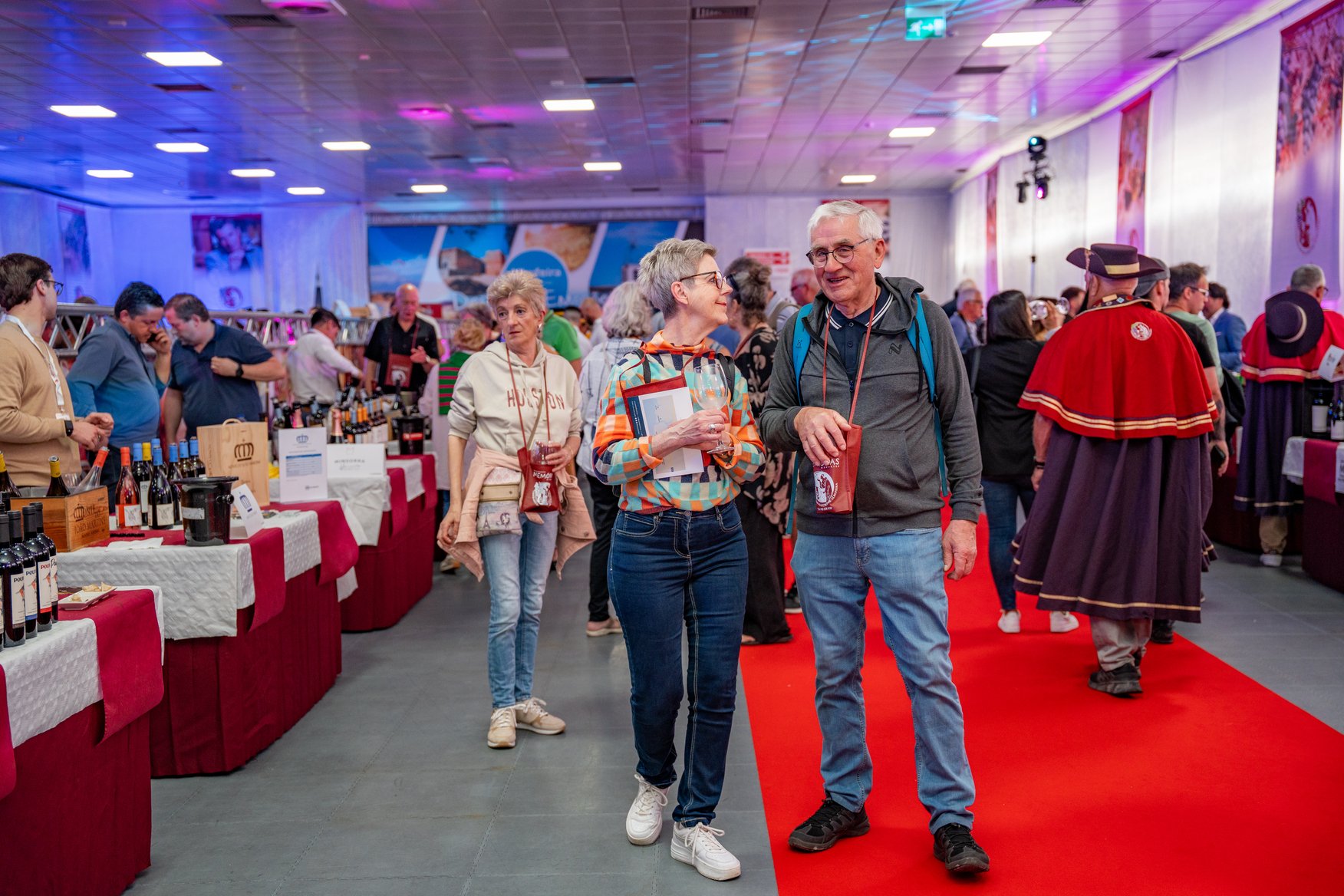 Pessoas conversando em uma feira de vinhos. Mesas com garrafas de vinho ao fundo, ambiente com iluminação colorida e decoração temática.
