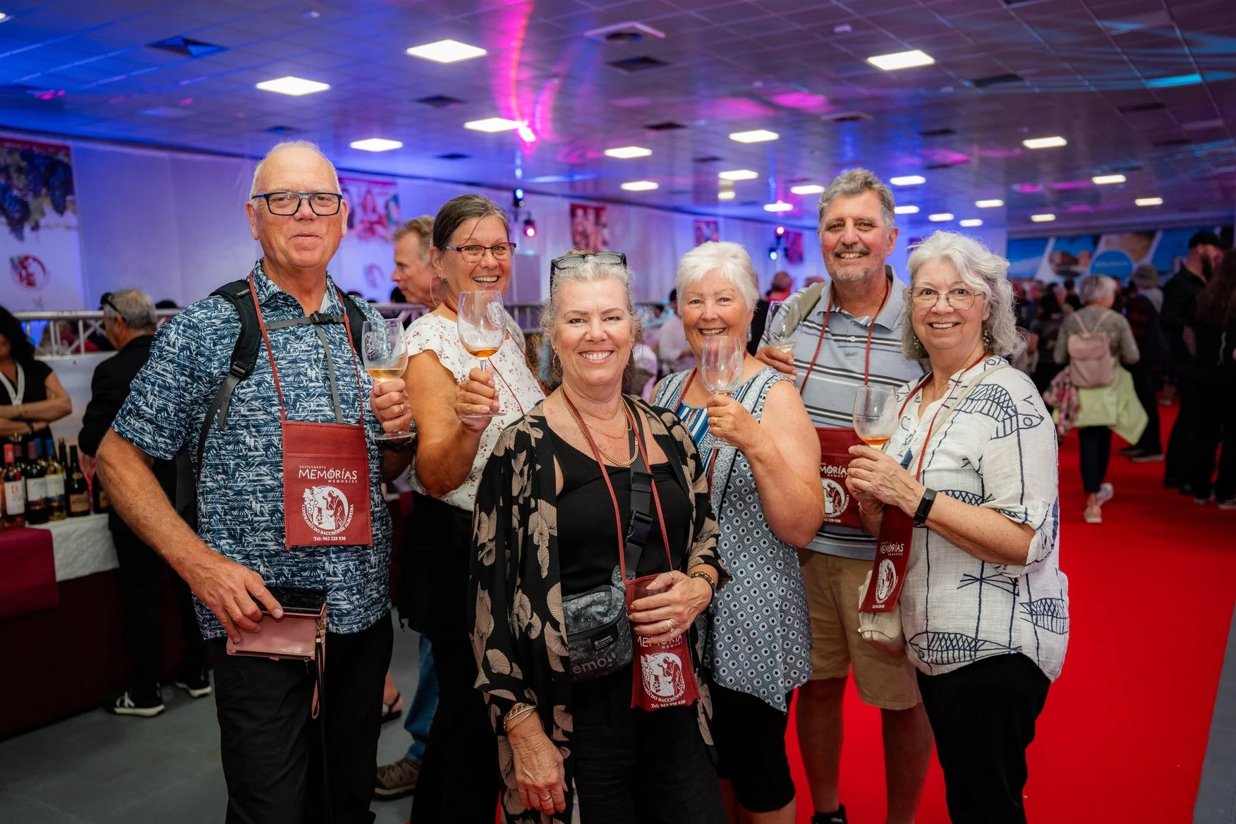 Grupo de idosos sorrindo na feira de vinhos, segurando taças de vinho em ambiente com luzes coloridas e decoração de evento.