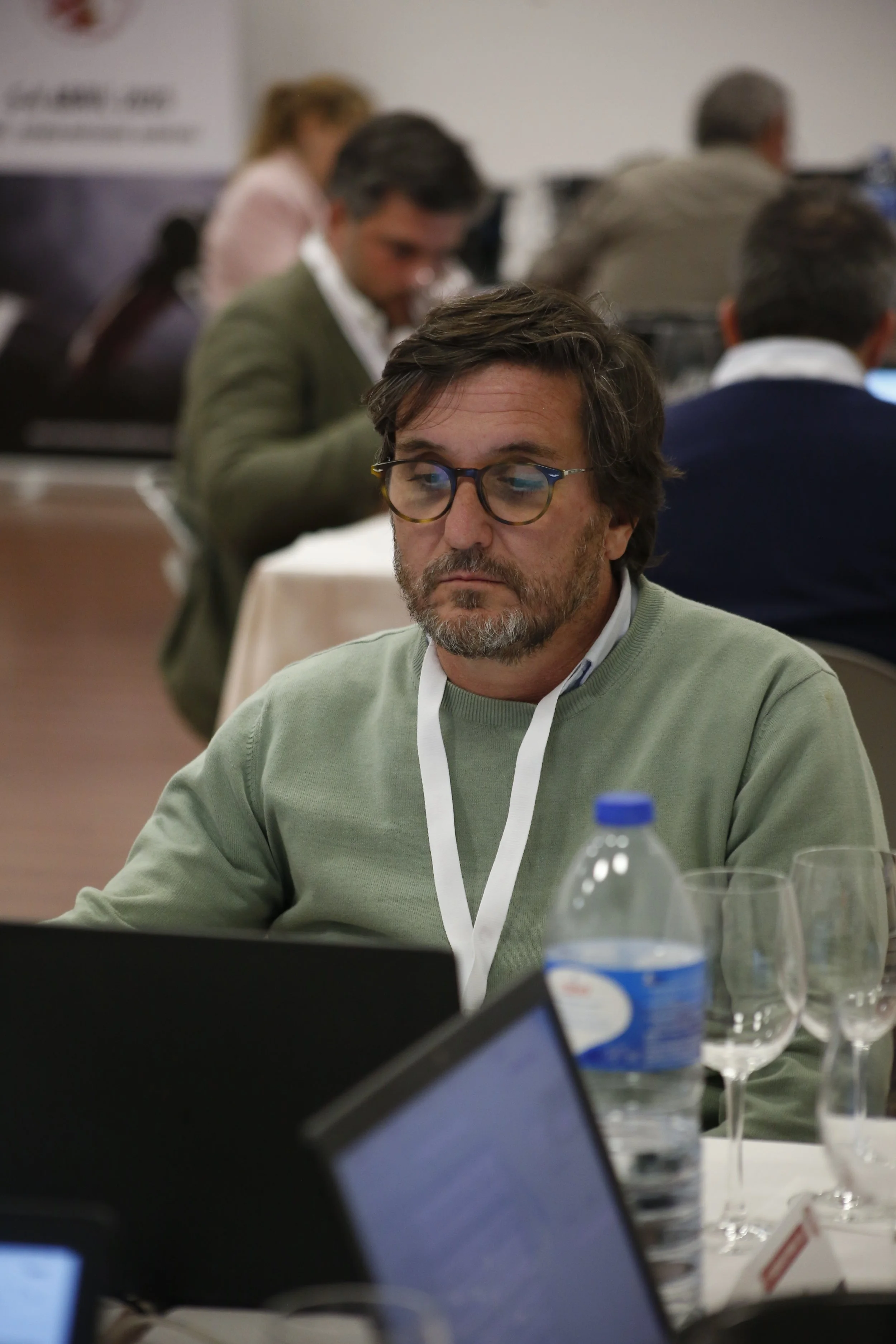Homem com barba e óculos sentado em uma mesa de conferência usando suéter verde, concentrado em um computador, com garrafas de água e taças ao redor, em ambiente de reunião.