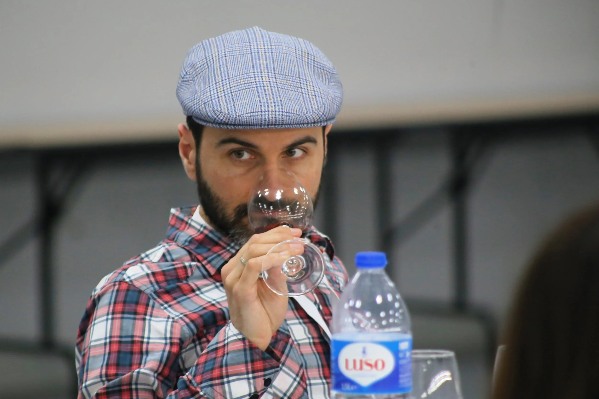 Homem segurando taça de vinho, usando chapéu estilo boina, camisa xadrez, ao lado de garrafa de água