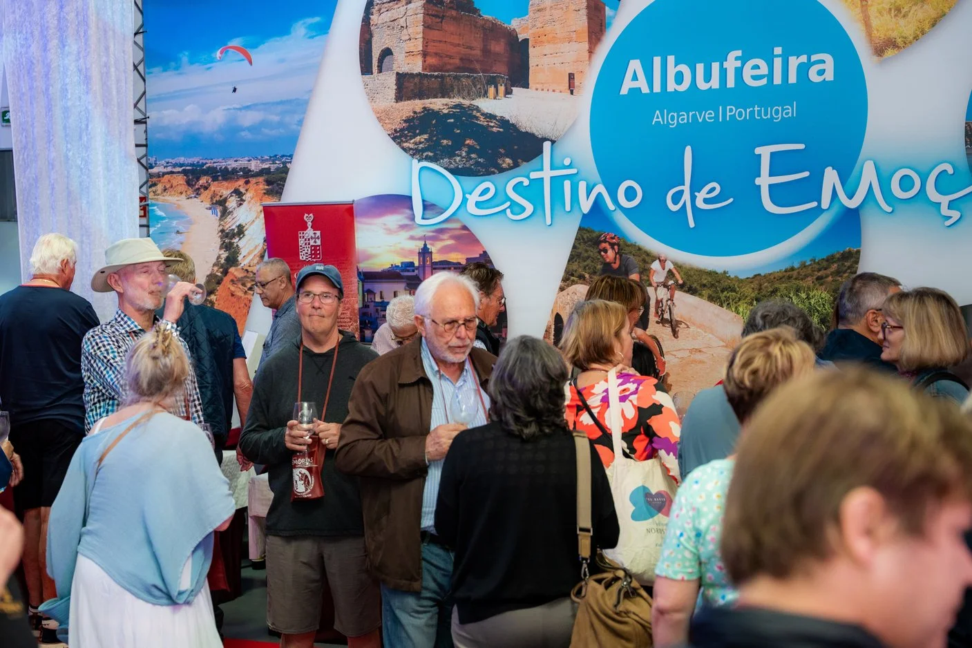 Pessoas em uma feira de turismo em Albufeira, Algarve, Portugal, com uma grande placa de fundo que diz 'Albufeira, Algarve Portugal, Destino de Emoção' e fotos das atrações locais.