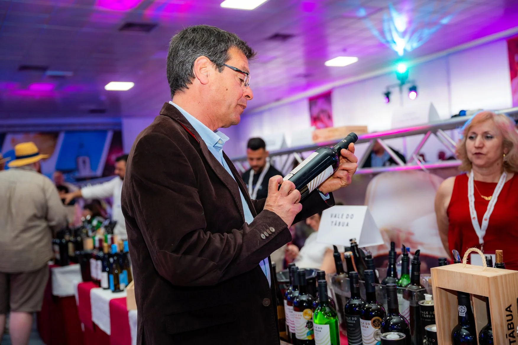 Homem olhando uma garrafa de vinho em uma feira com várias outras garrafas na mesa, ao fundo pessoas e um ambiente com iluminação colorida.