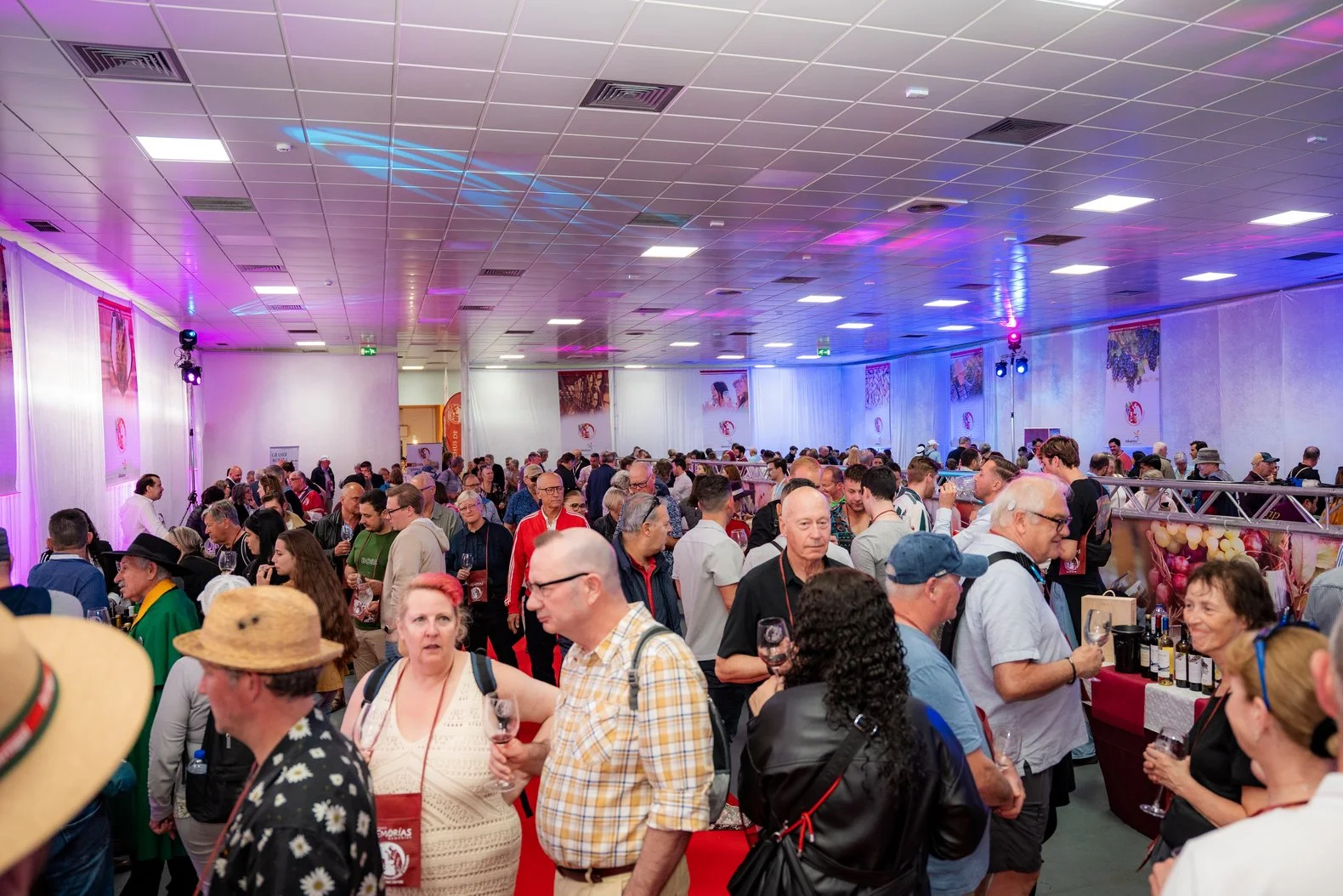 Imagem de uma feira ou evento com muitas pessoas, alguns provando vinhos, em um ambiente com decoração de luzes coloridas e banners nas paredes.