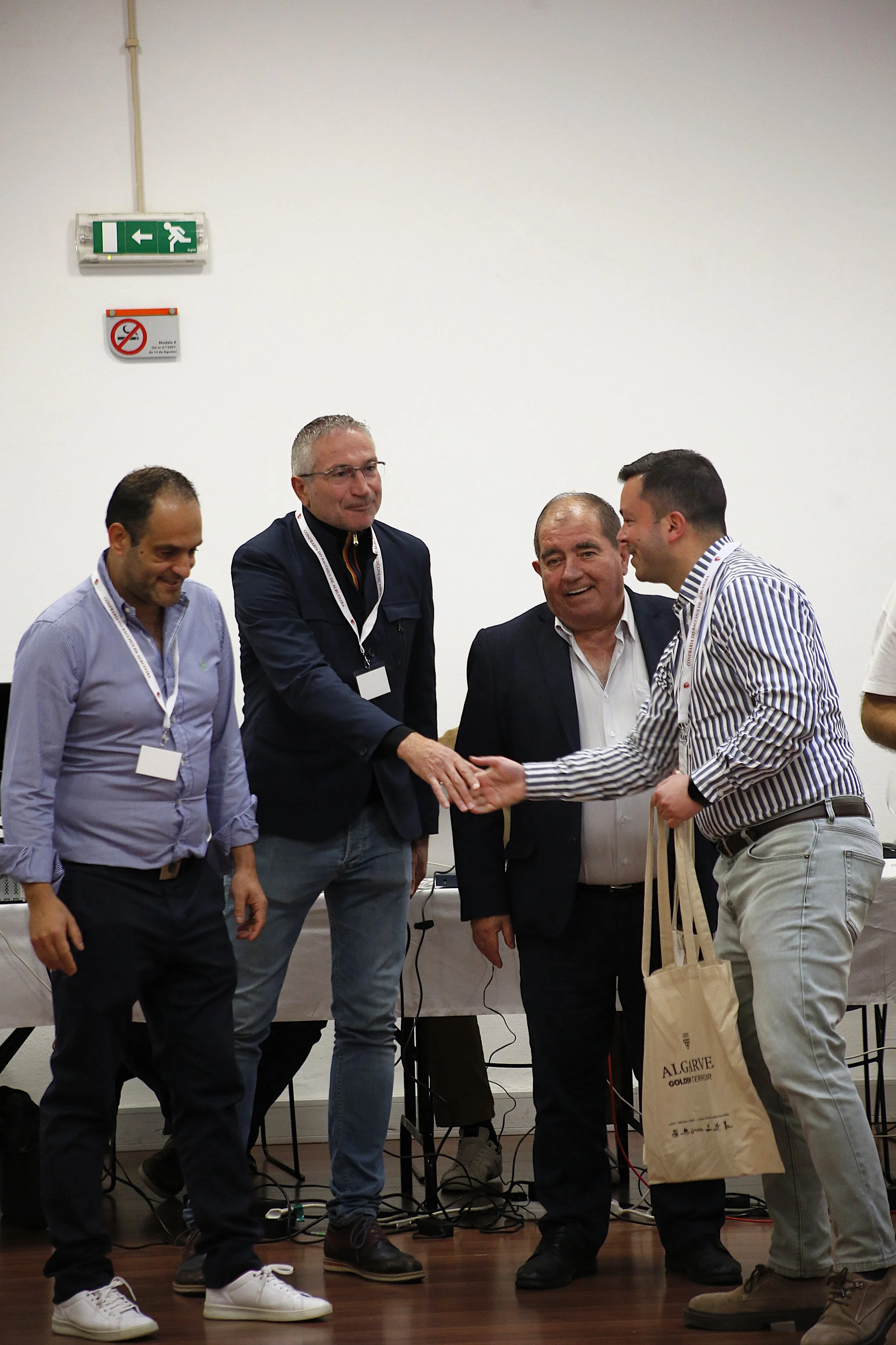 Homem recebendo prêmio ou reconhecimento em cerimônia, com outras pessoas ao fundo, em ambiente interno.