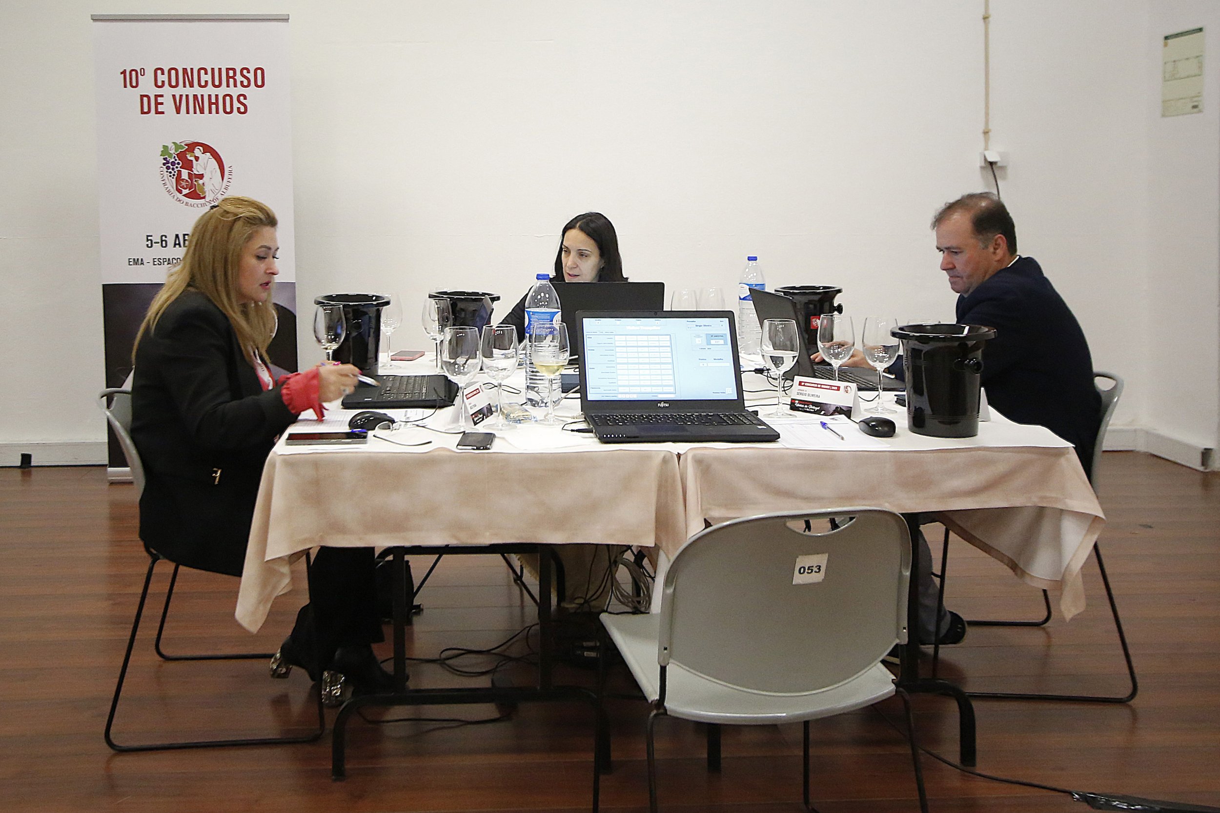 Três pessoas sentadas em uma mesa de julgamento de vinhos com laptops, copos, garrafas d'água, e equipamentos de degustação, em uma sala com uma faixa promocional ao fundo.