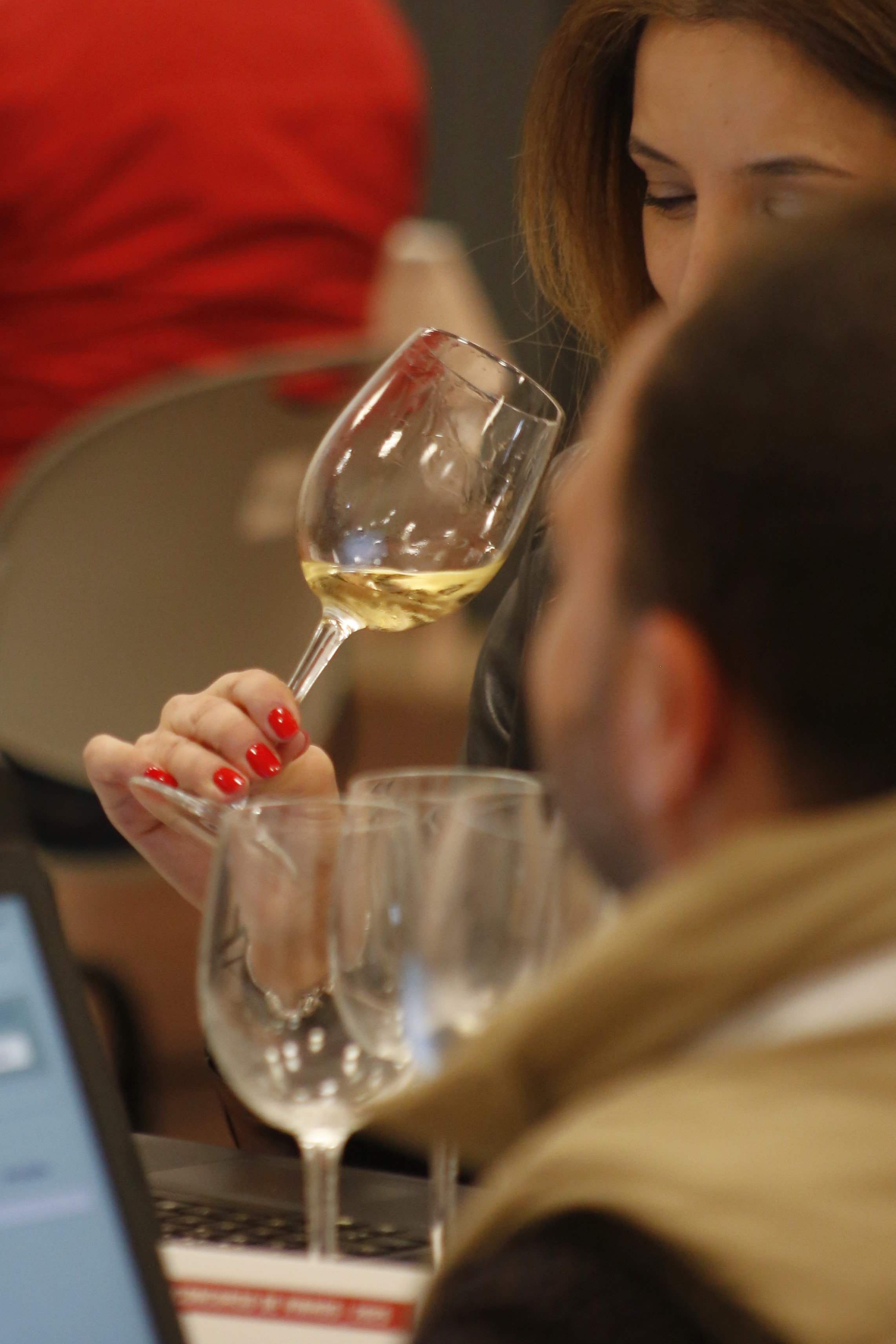 Pessoa segurando taça de vinho branco em um ambiente social, com outras taças e pessoas ao fundo.