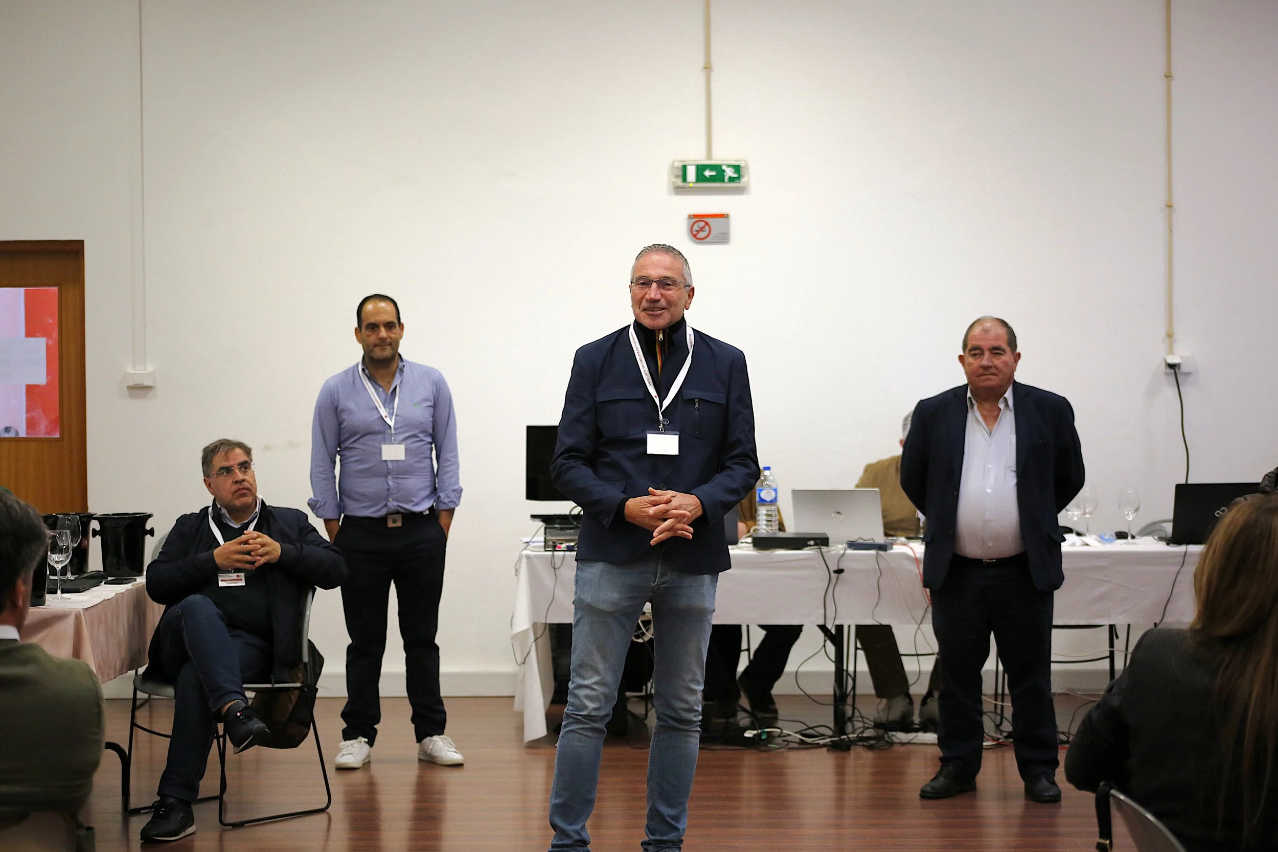 Homem sorridente falando em uma sala de reunião com várias pessoas ao redor, alguns sentados e outros em pé, usando crachá. Ao fundo, mesa com computadores, garrafas de água e taças, parede branca e sinal de emergência.