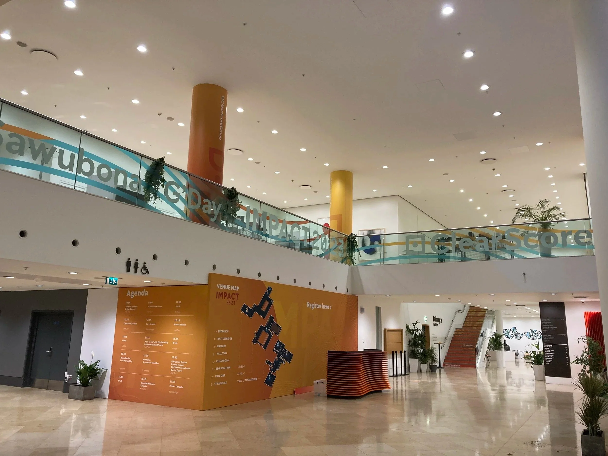 Interior of a modern shopping mall or conference center with a large open area, potted plants, informational signs, and a staircase leading to an upper level. The upper level has a glass railing with text references, and decorative greenery.