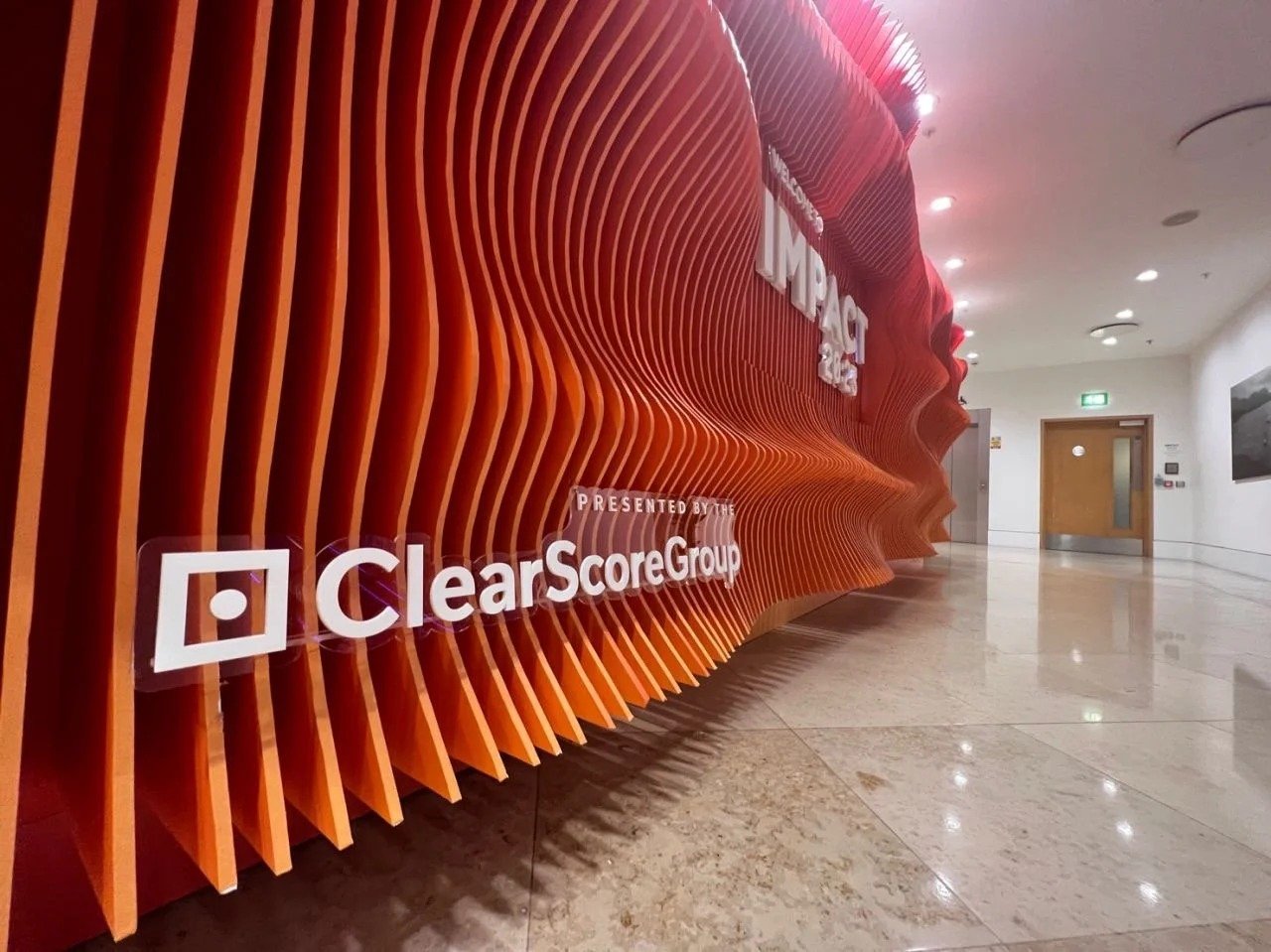 Red modern wall sculpture with company logos including ClearScoreGroup and Impac, in a corporate lobby hallway.