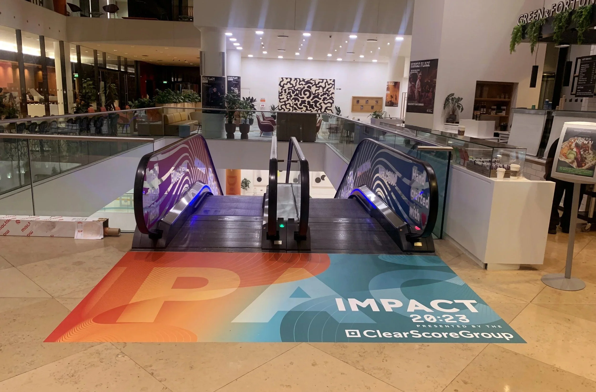 Empty escalators in a shopping mall with a large banner on the floor advertising an event called IMPACT at 20:23, presented by ClearScoreGroup.
