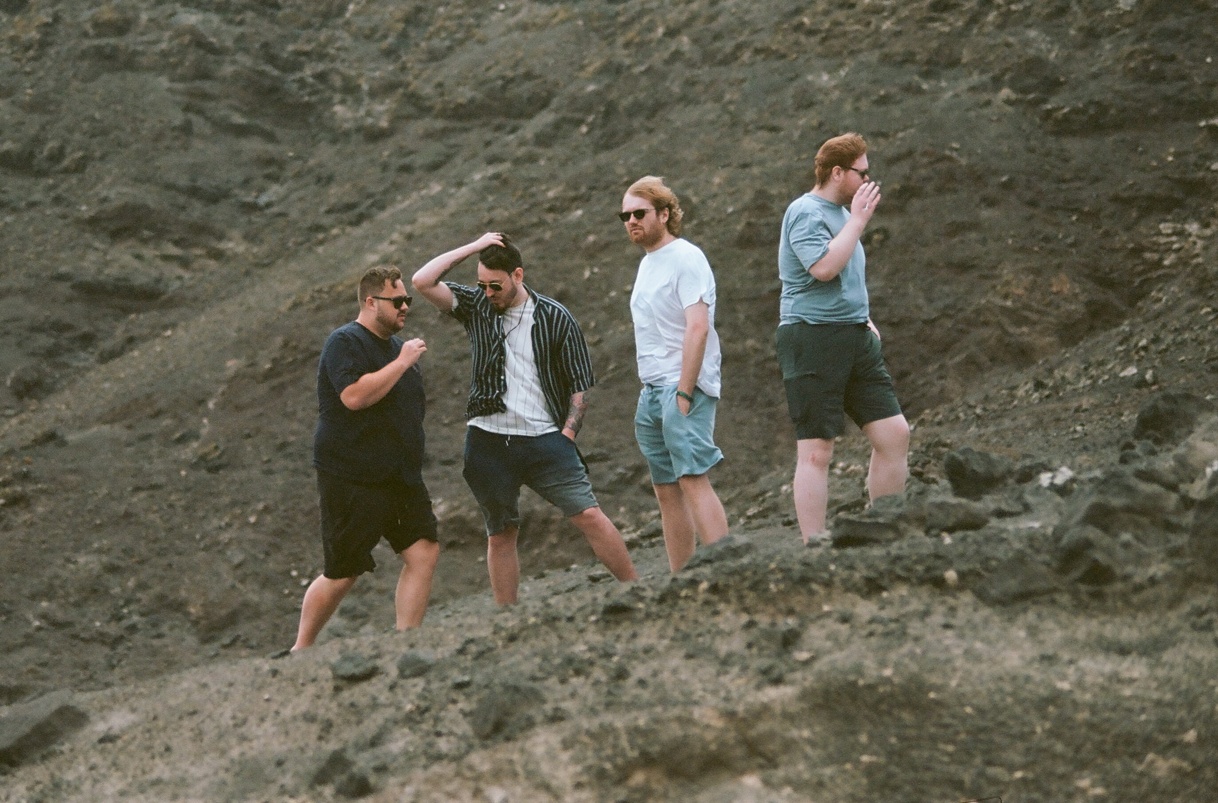 Four men standing on rocky terrain, maybe at a beach or volcanic area, wearing casual shorts and t-shirts, some with sunglasses.