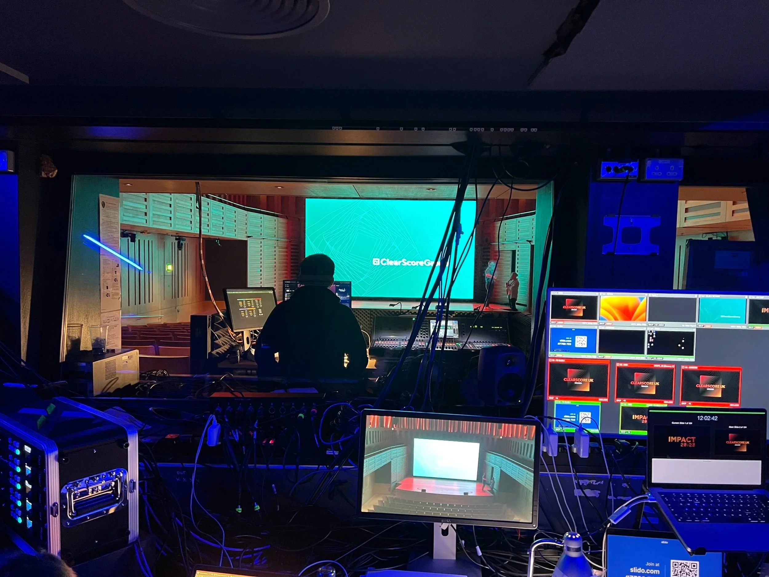 View of a production control room looking into an auditorium stage with a large screen displaying a blue geometric background and the words 'ClearScore Group.' The control room has multiple monitors, equipment, and a technician working at the console