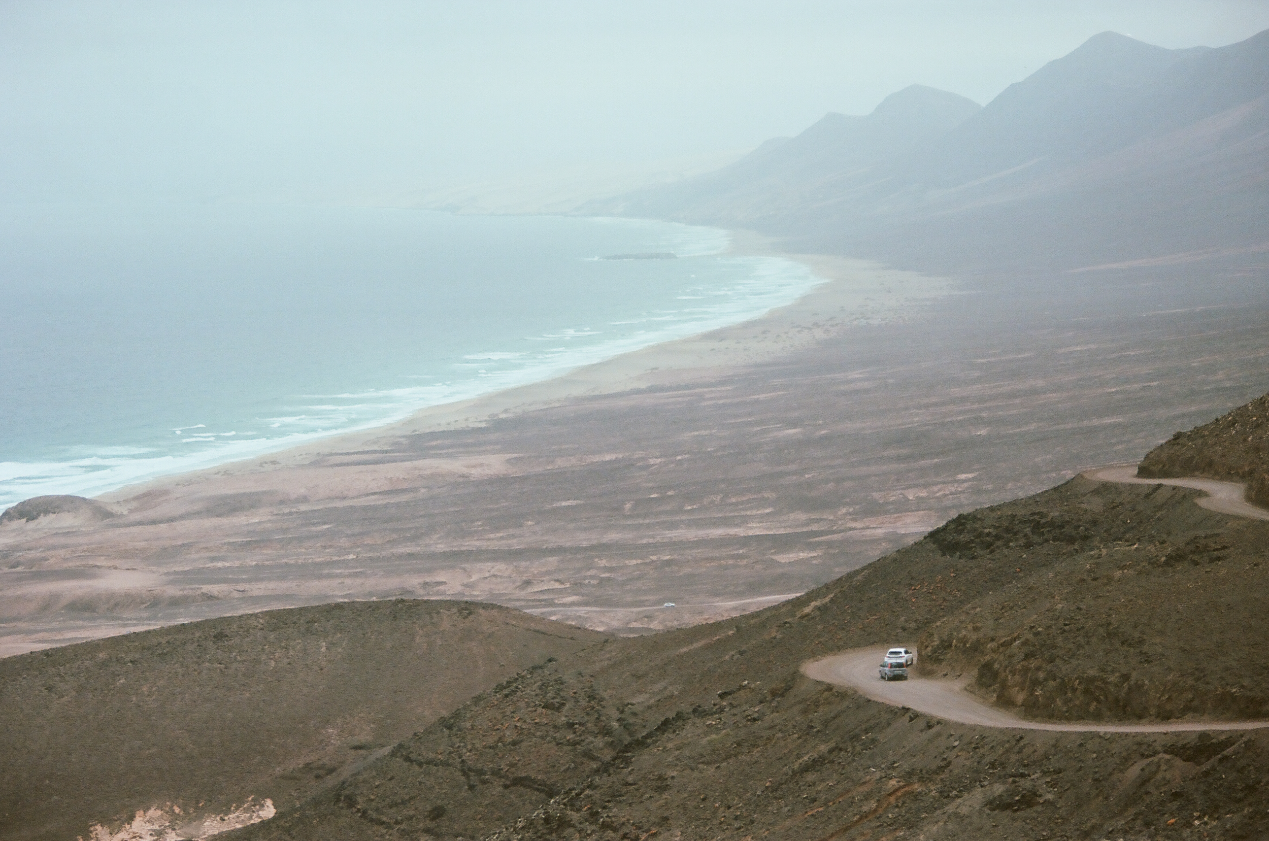 Winding dirt road on mountainside overlooking a coastal beach and distant mountains, with two white vehicles traveling along the road.