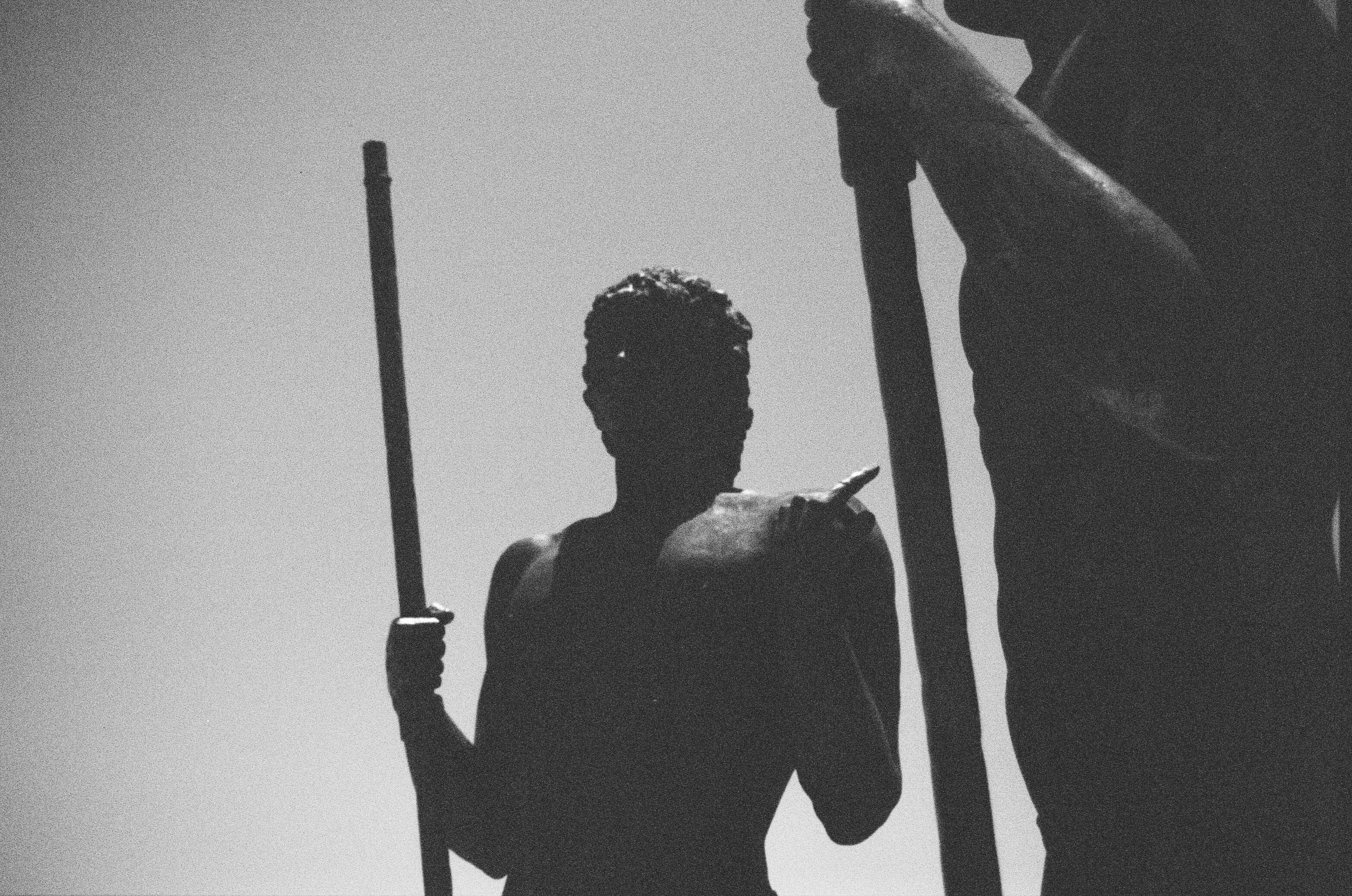 Silhouette of a person holding a pole or stick, with another person partially visible on the right side, against a clear sky.