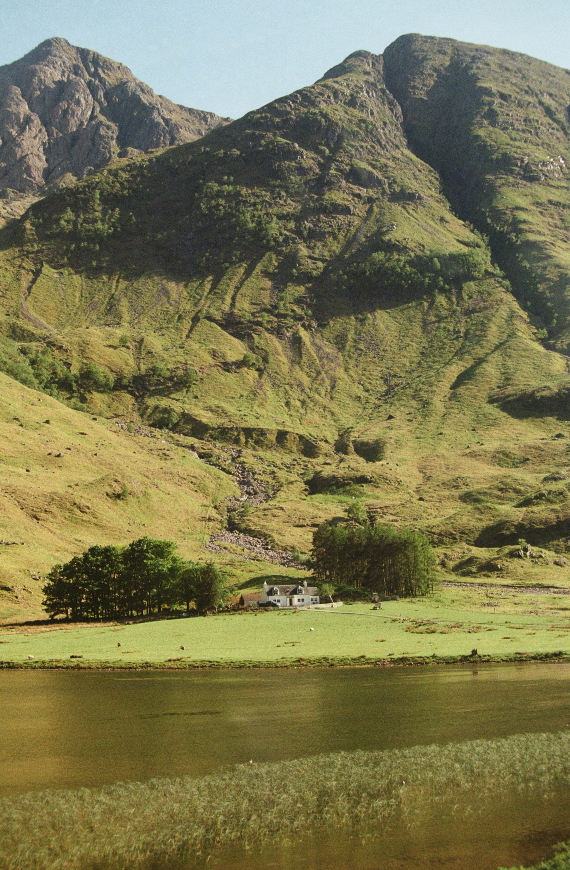 Lush green mountains and hills with a small white house and trees by a body of water.