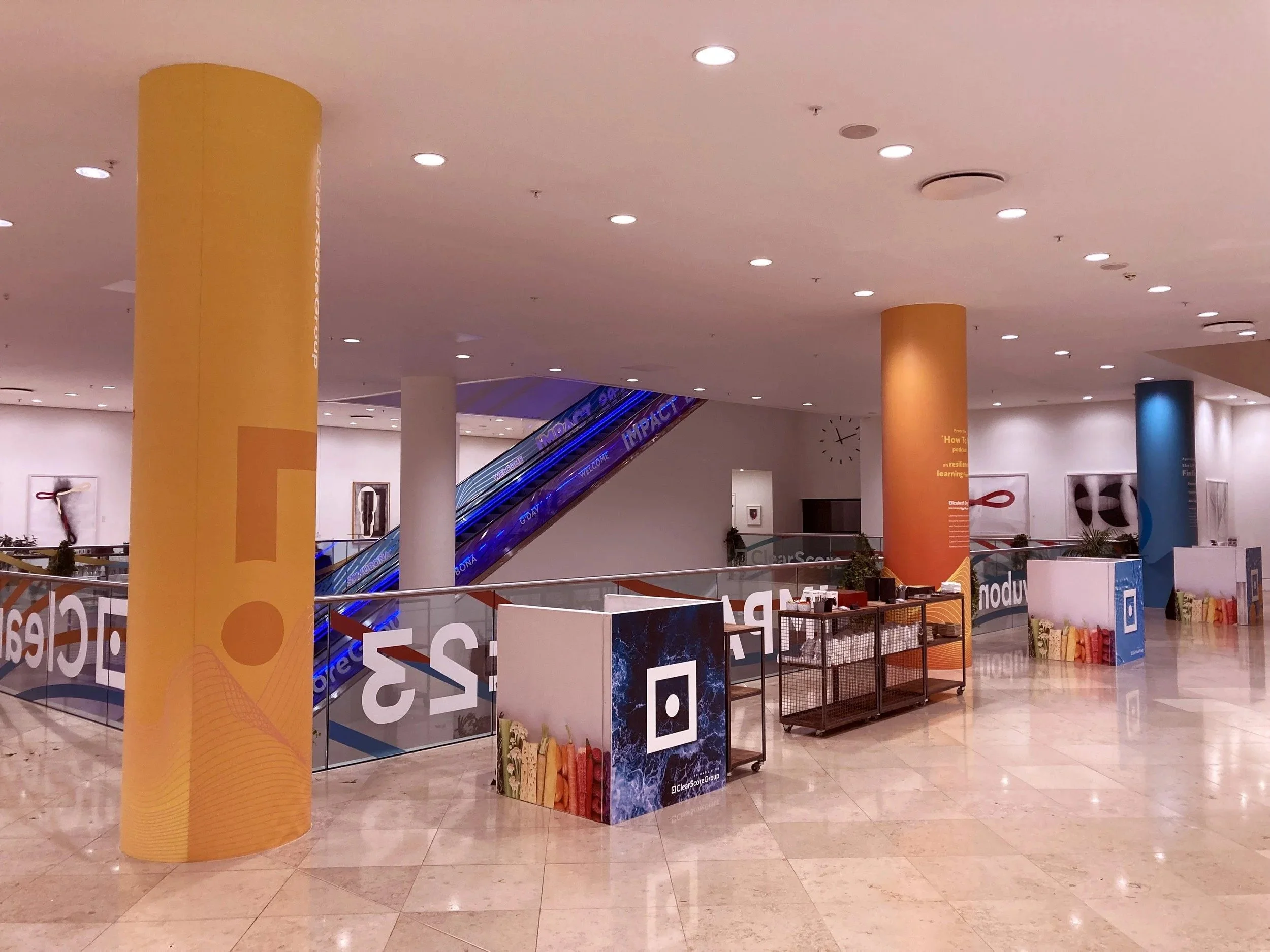 Empty shopping mall area with large colorful pillars, escalator in the background, store displays, and signage.