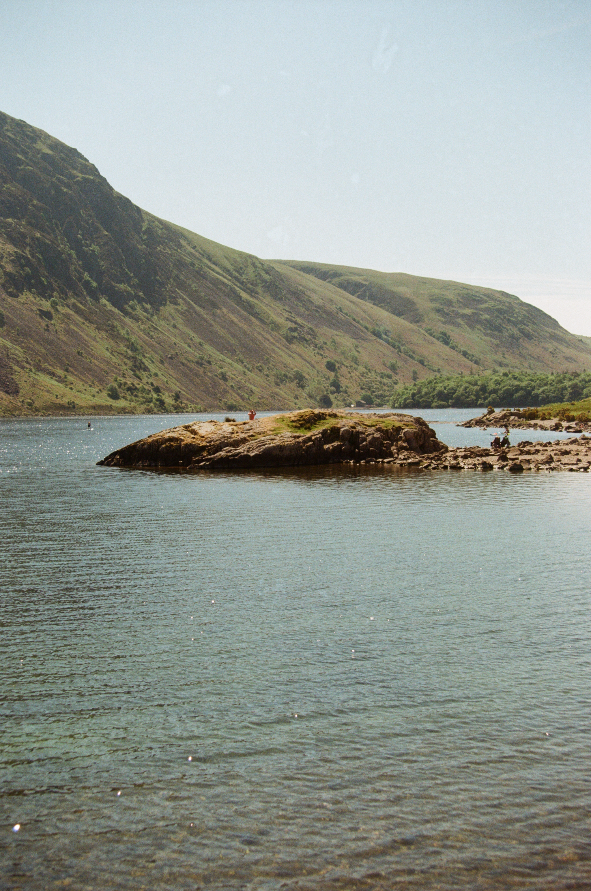 A serene lake surrounded by green hills and mountains, with a rocky island or peninsula in the foreground and a person fishing on the shore.