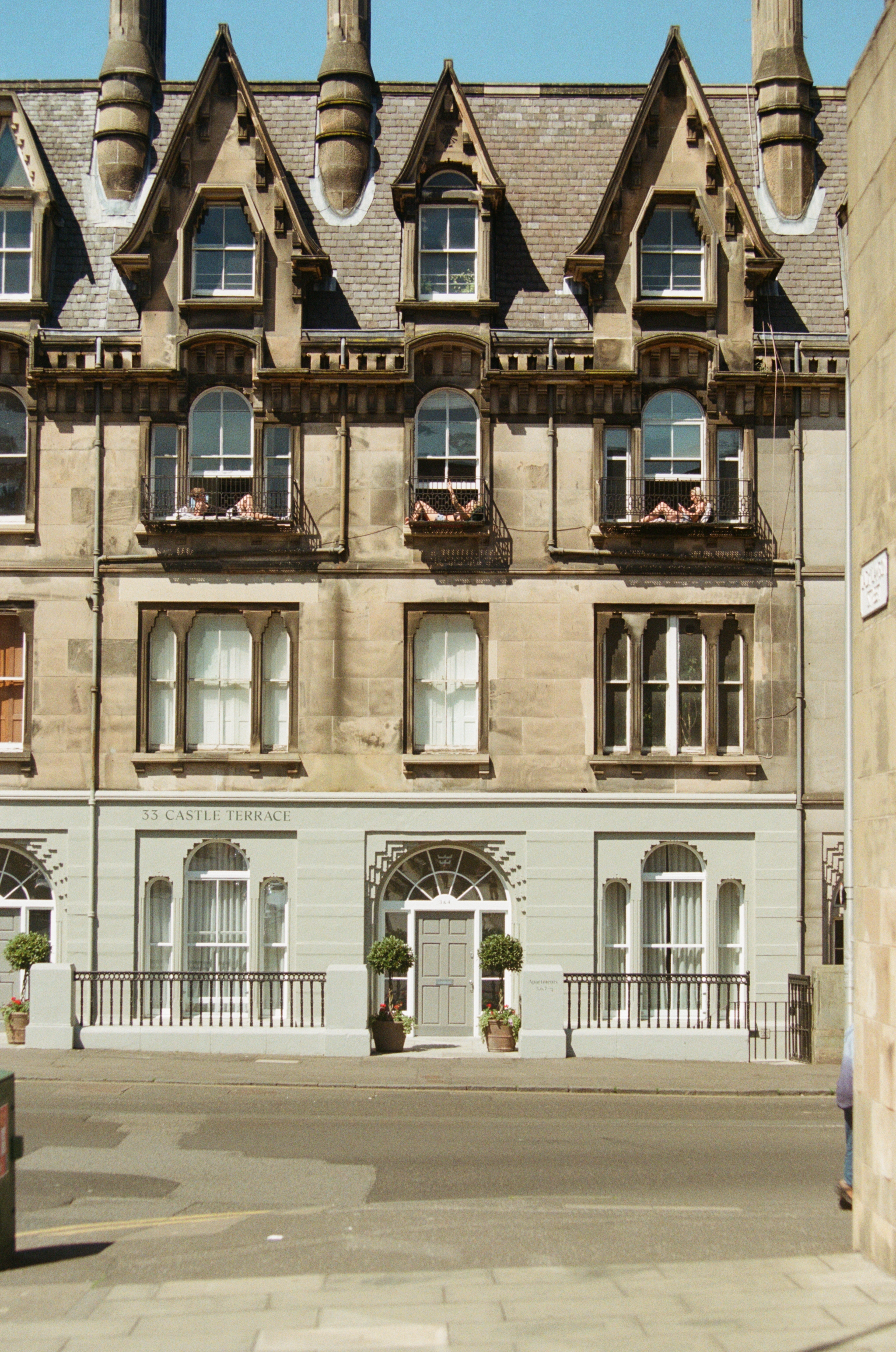 A historic multi-story building with Victorian architecture, featuring large windows, ornate details, and balconies with people lounging. The building is marked with the address 53 Castle Terrace and has potted plants at the entrance.