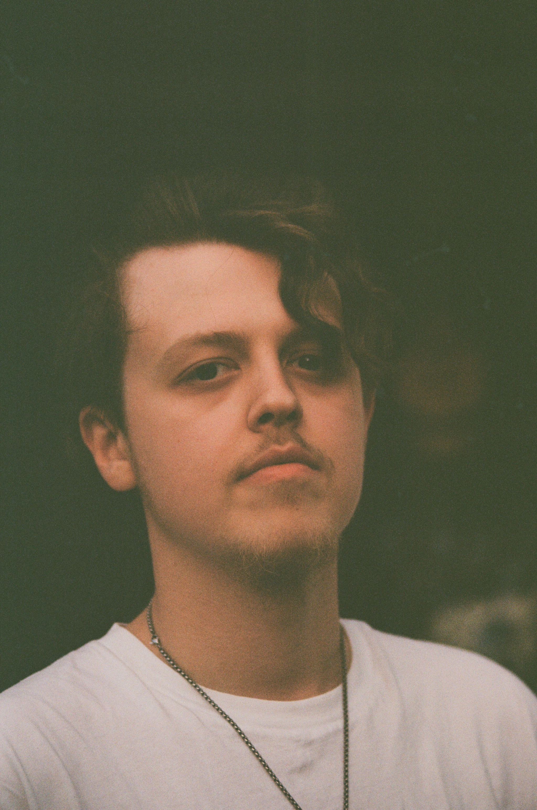 A young man with brown curly hair and a slight beard, wearing a white shirt and a chain necklace, is looking at the camera with a neutral expression. The background is dark and blurry.