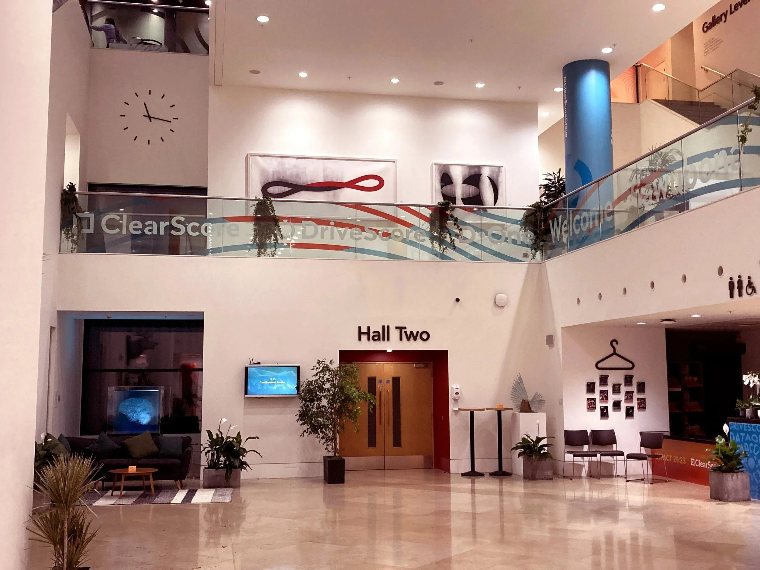 Interior of a modern building with a sign that reads 'Hall Two' on a white wall, seating area with chairs, plants, and a wall display of photos, staircase on the right, and a blue column on the right side.