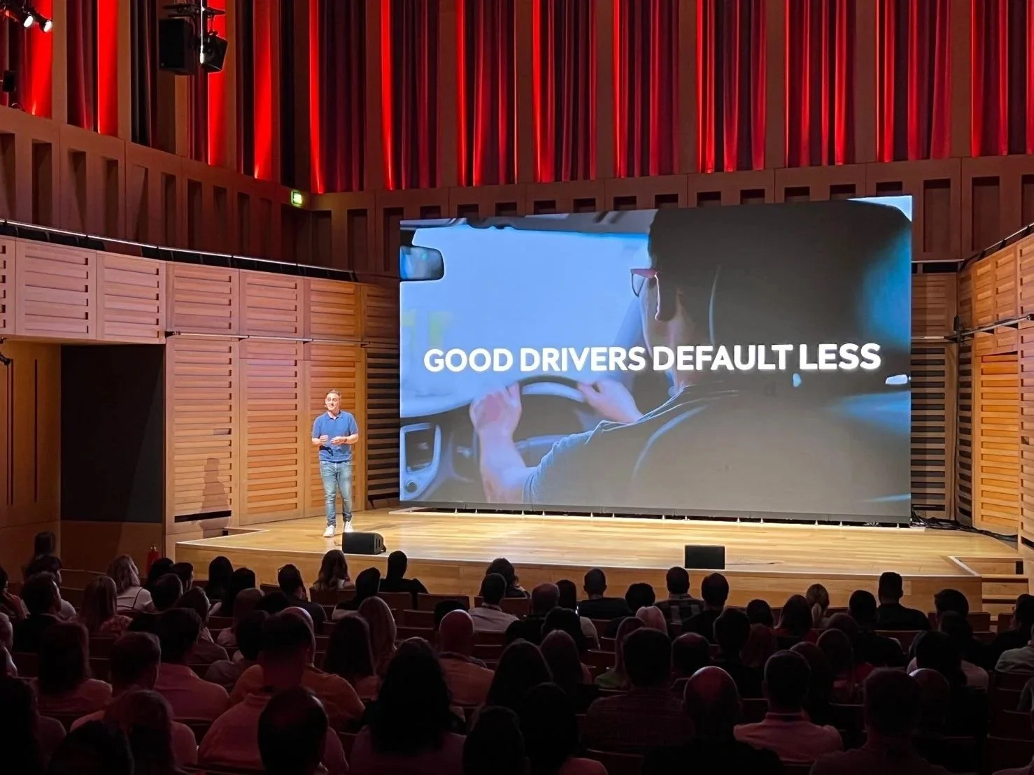 A speaker presenting on stage in front of a large audience with a slide showing a woman driving a car and the text "Good drivers default less."