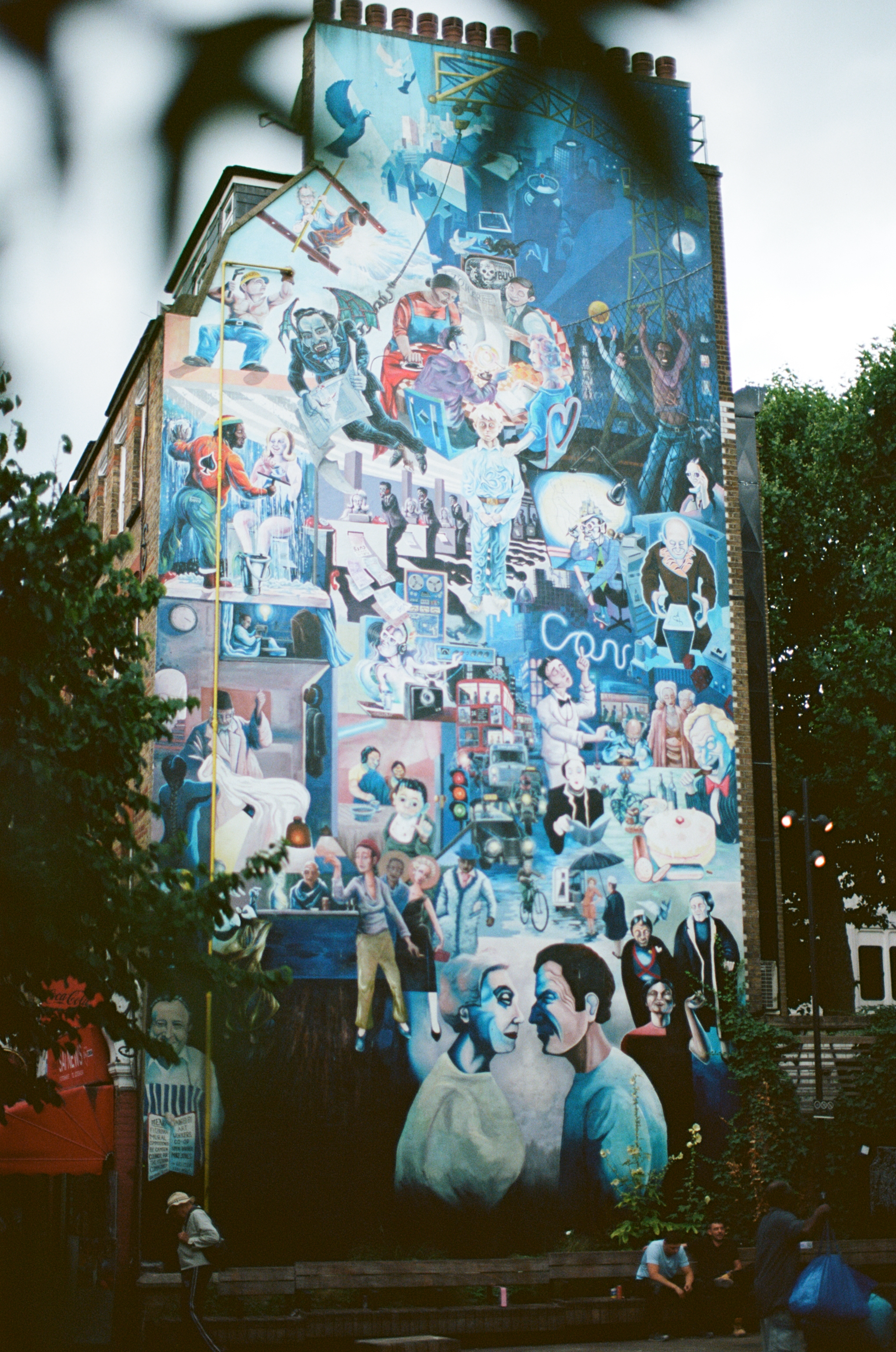 A colorful mural on the side of a building depicting various scenes of people engaging in different activities, including art, science, and everyday life, with some large faces at the bottom foreground and trees surrounding the building.