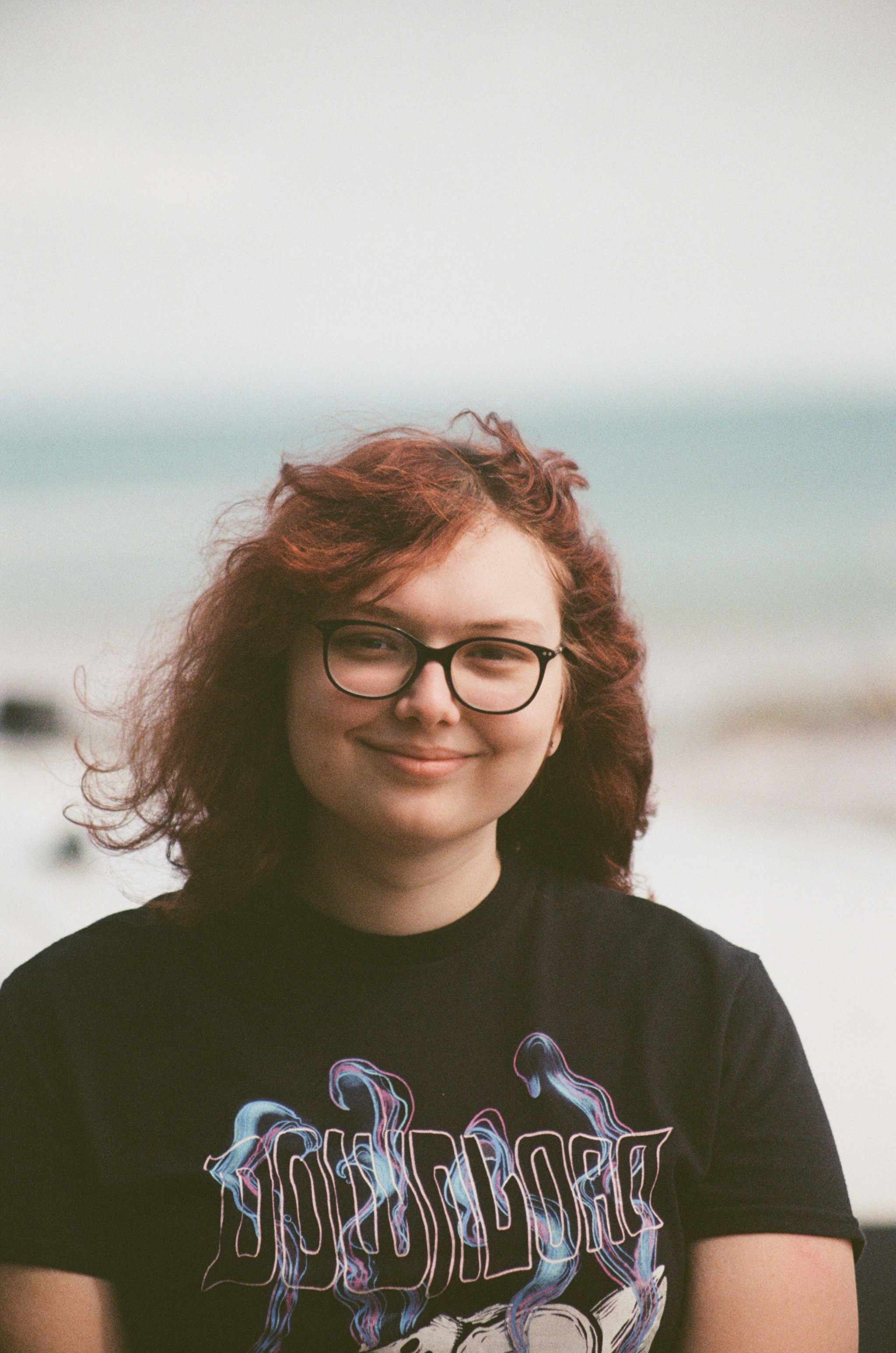 Young woman with curly red hair and glasses smiling at the beach, wearing a black graphic t-shirt.
