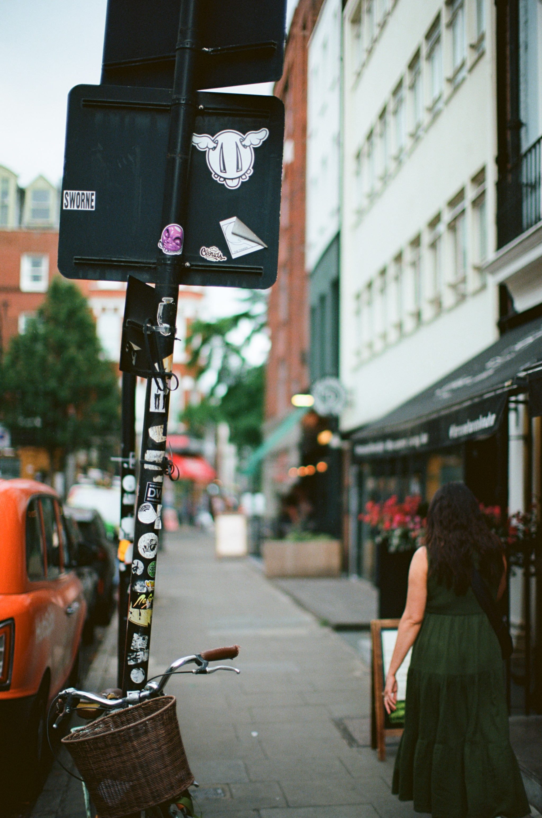City street scene with a person walking away, a bicycle with a wicker basket, parked cars, storefronts, and a pole with various stickers and decals.