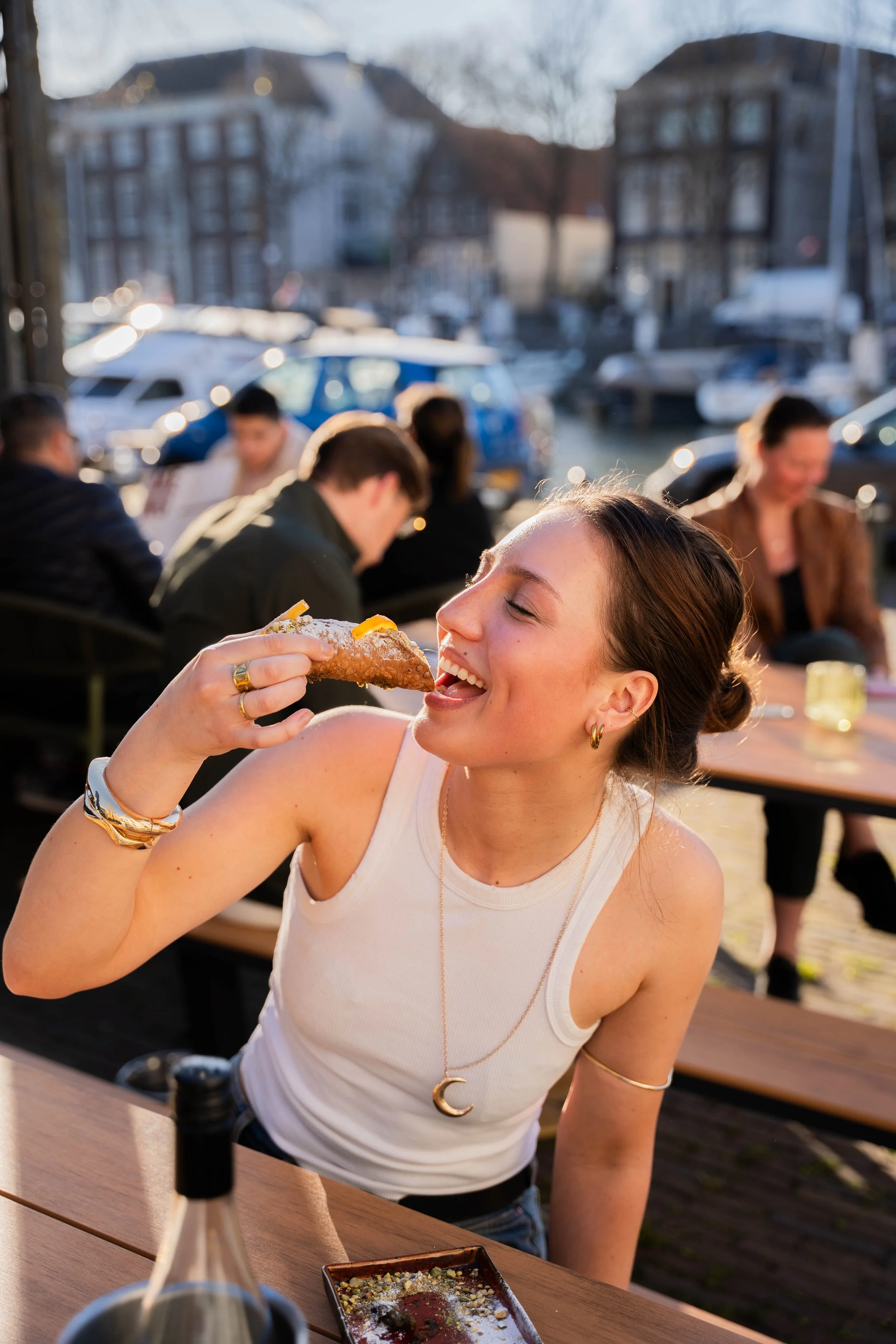 A young woman smiling and about to eat a slice of pizza outdoor at a restaurant with a marina and boats in the background, during sunset.