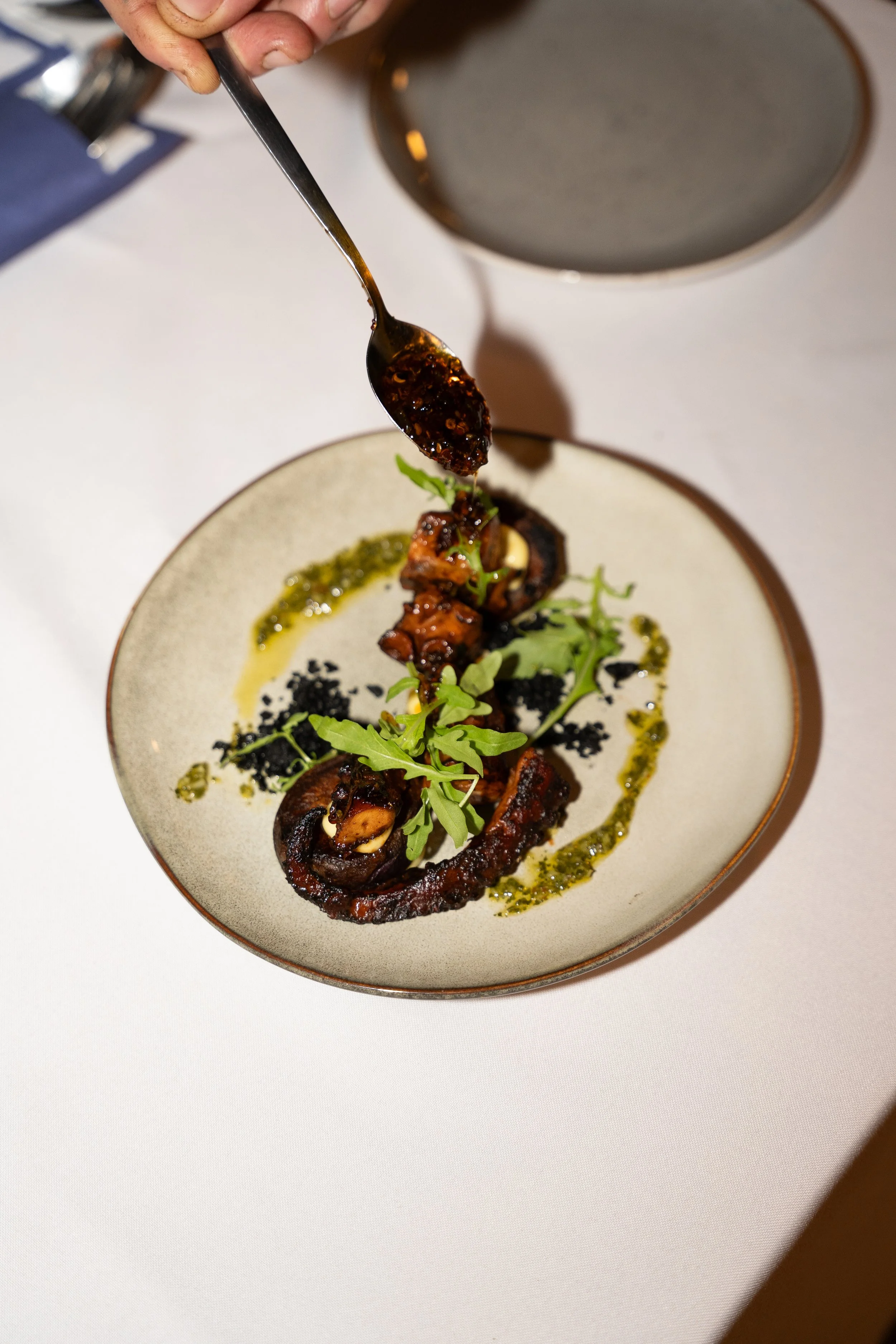 Upscale image of a plated gourmet dish featuring grilled octopus, garnished with leafy greens, accompanied by sauces and herbs, on a white table.