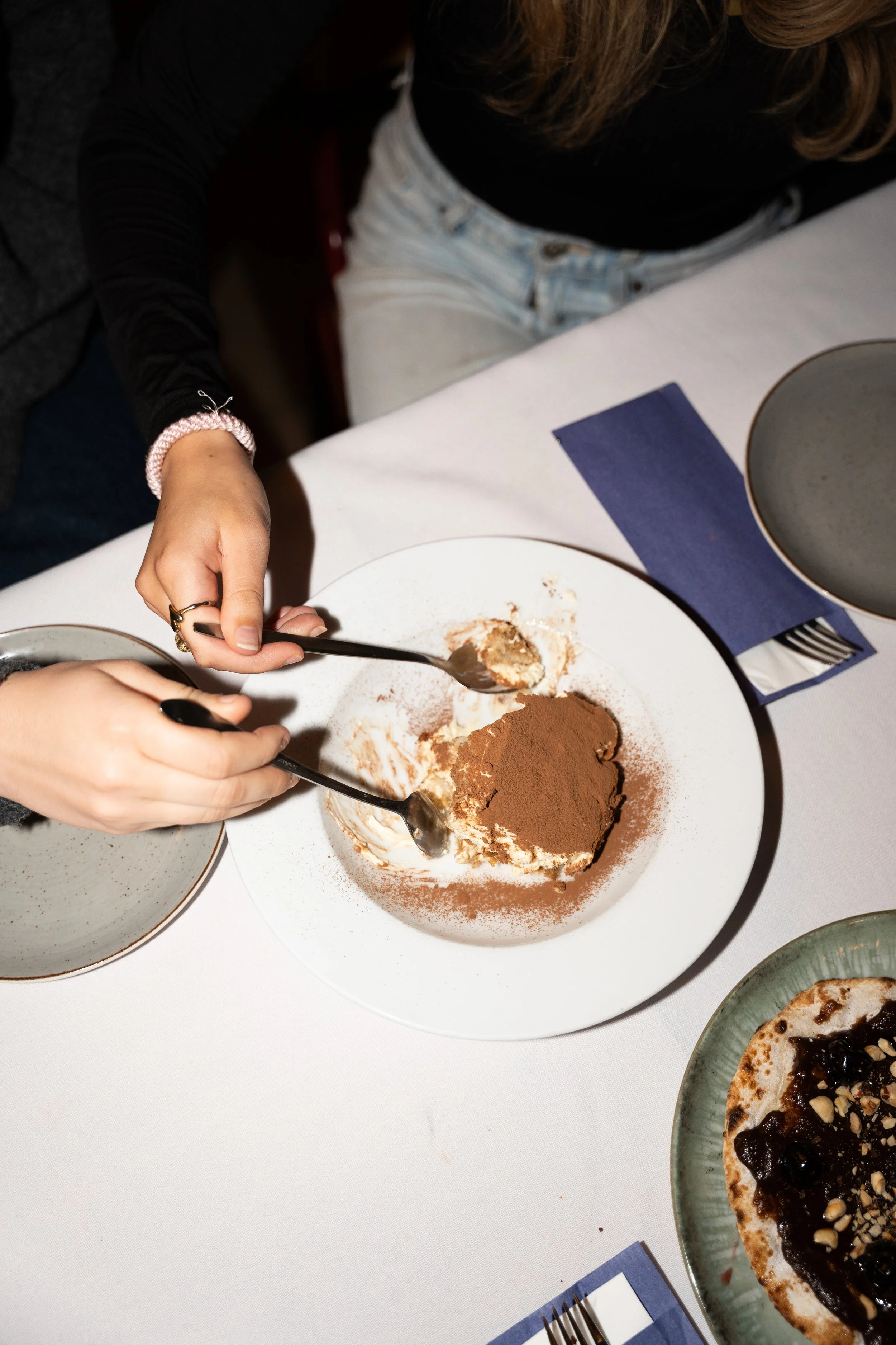 Person scooping tiramisu dessert with two spoons on a white plate at a table with bowls, napkins, and a pizza.