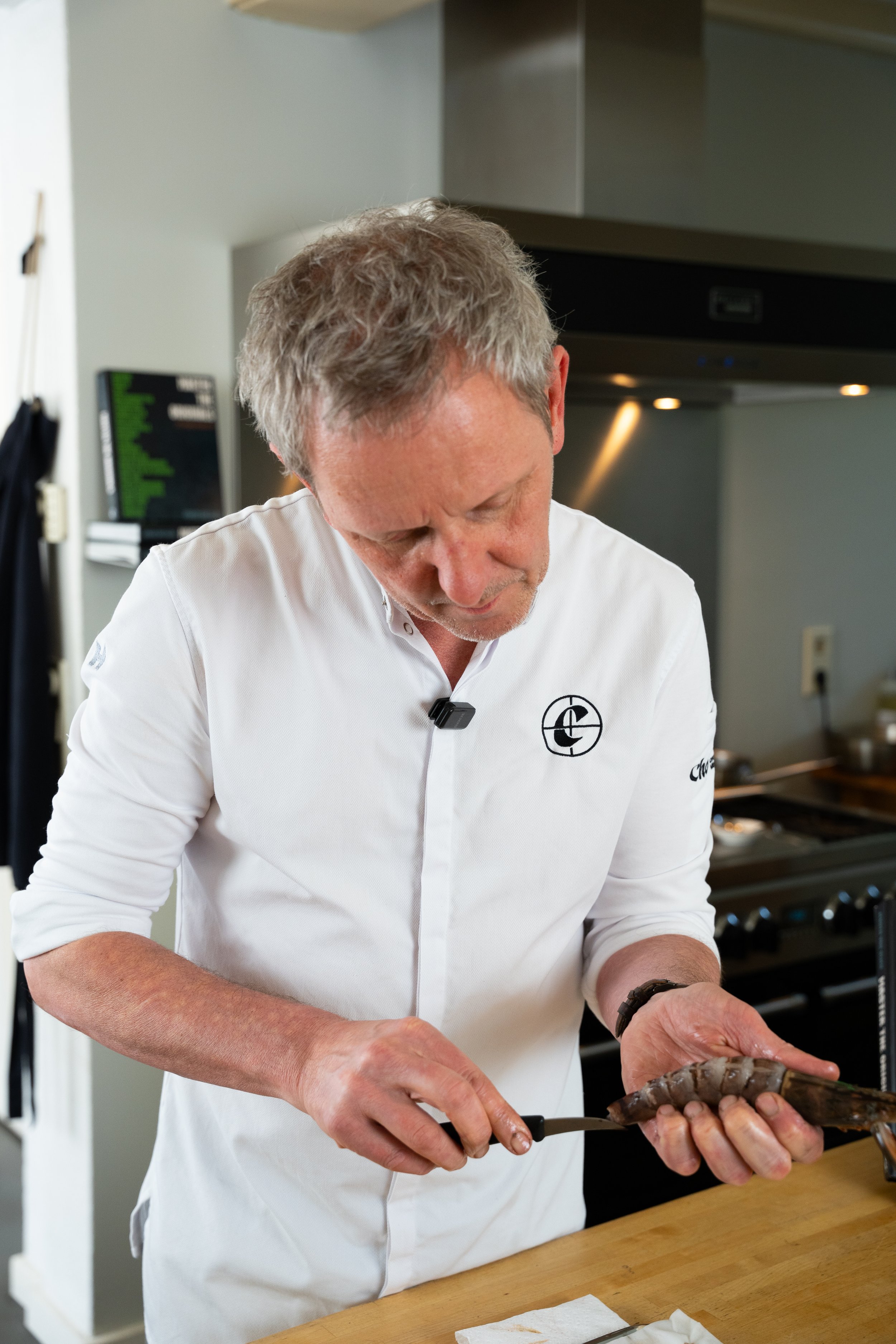 A man with gray hair wearing a white chef's coat with a logo, preparing food in a kitchen.