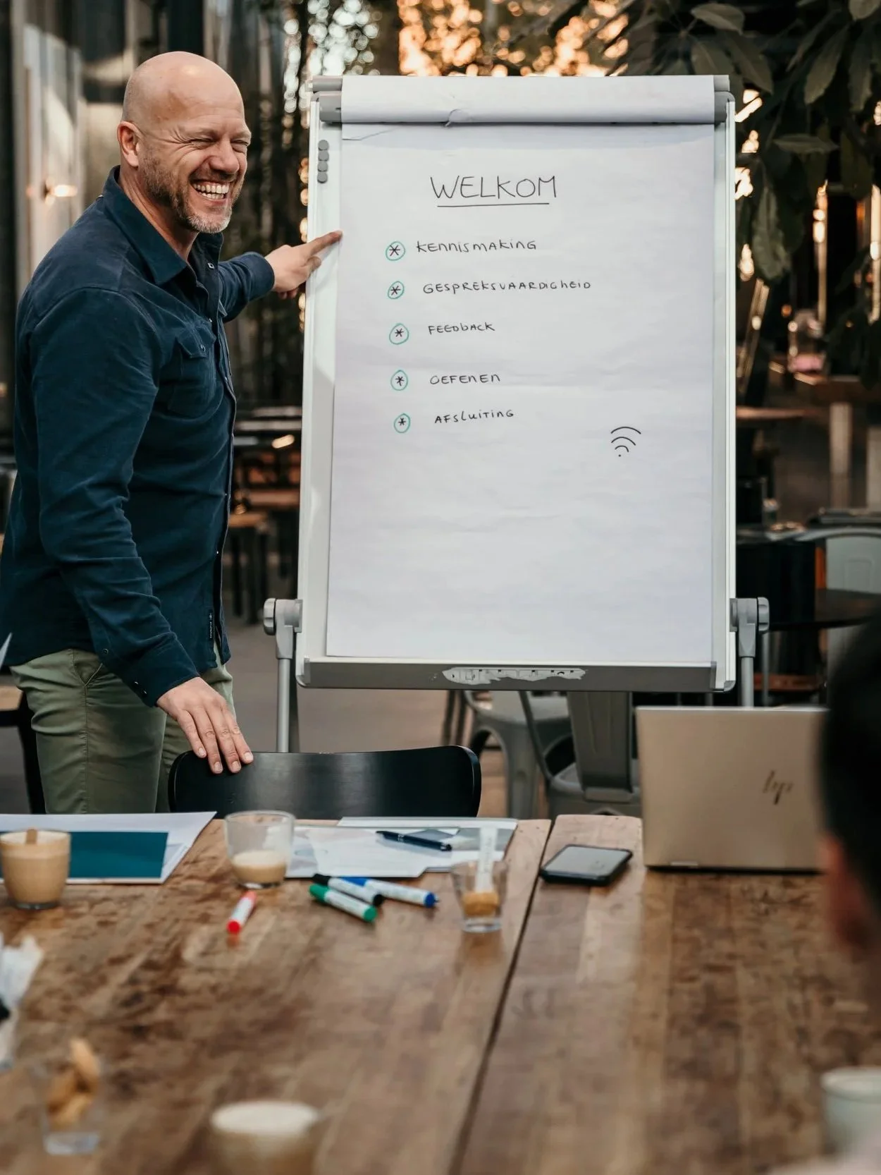 A man is smiling and pointing to a whiteboard with a list of items, including 'kenskmaking,' 'gespreksvaardigheid,' 'feedback,' 'oefenen,' and 'afsluiting.' The setting appears to be a meeting or workshop in a casual, outdoor environment with tables, papers, pens, and coffee cups in the foreground.