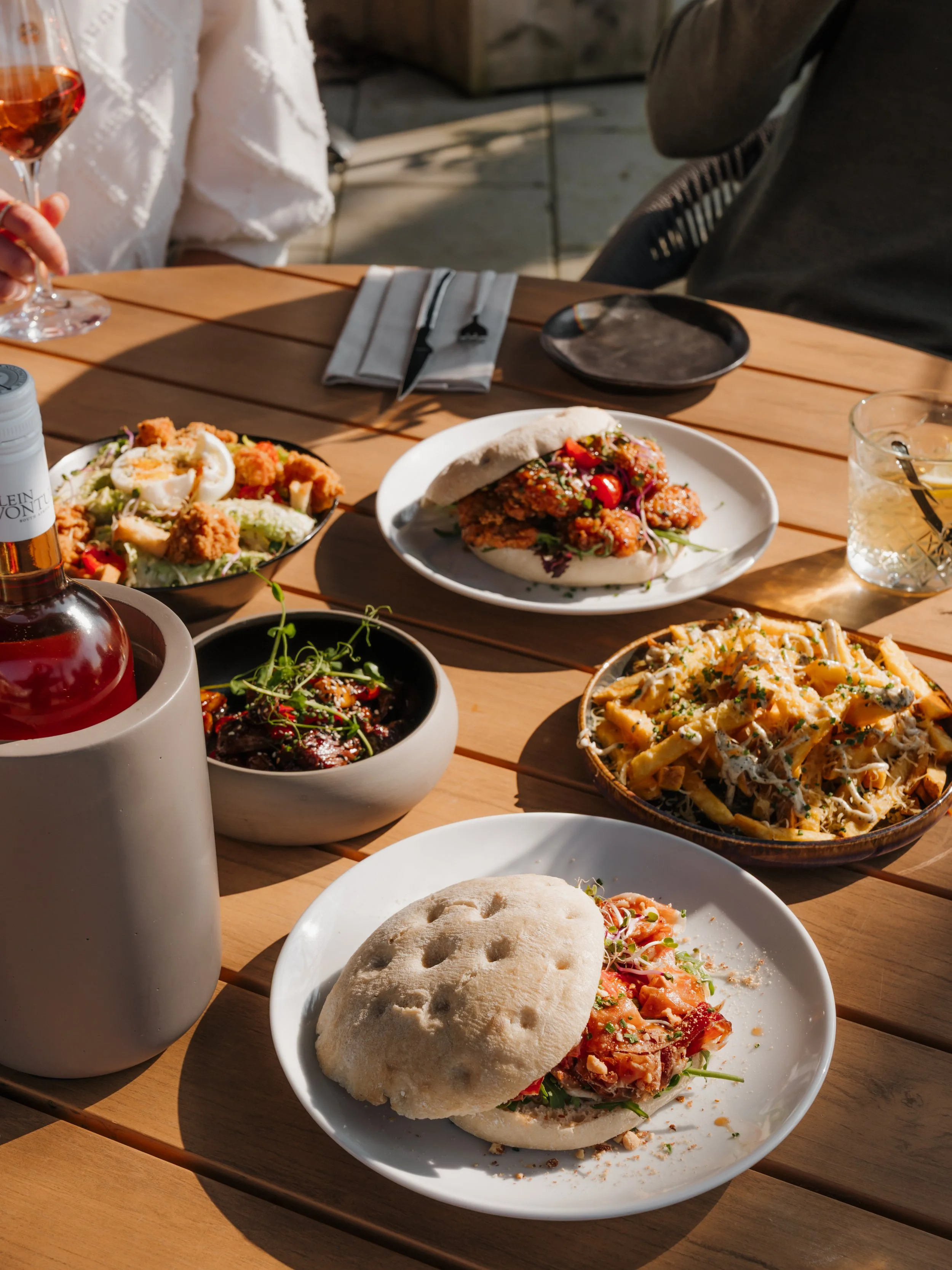 A table with various dishes including a bao bun with fried chicken and vegetables, pasta with sauce, a salad with greens and boiled egg, and a bowl of rice with vegetables, along with two glasses of drinks and a bottle of wine.