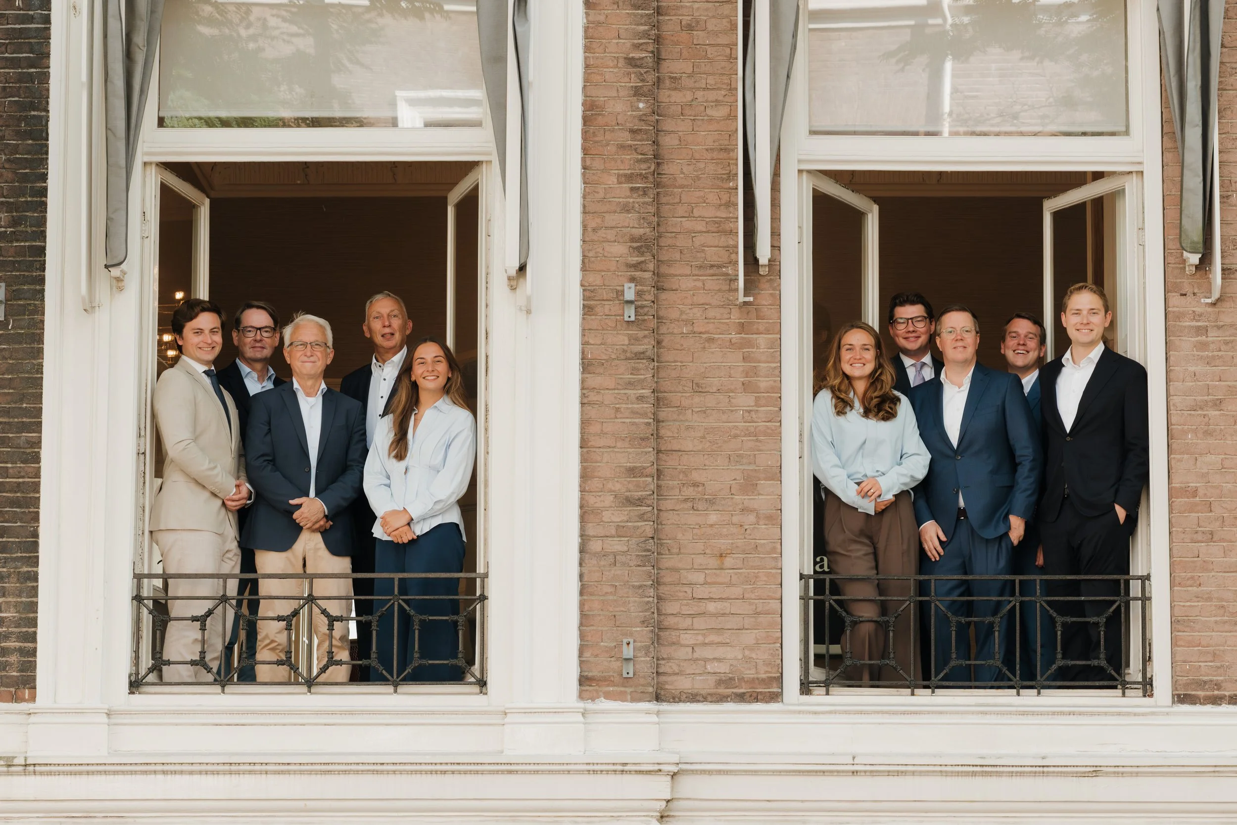 Tien mensen in zakelijke kleding staan op twee naast elkaar gelegen balkons van een gebouw, ieder achter een open raam. De groep bestaat uit zowel mannen als vrouwen, glimlachend en gekleed in formele kleding. Het gebouw heeft een bakstenen gevel en witte raamkozijnen.