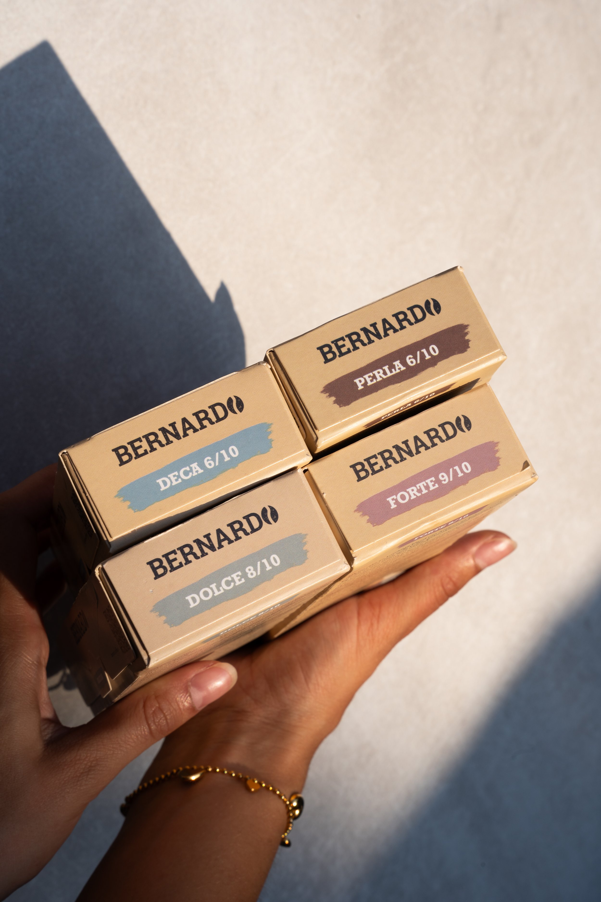 Person holding a stack of four Beauty product boxes with various shades and labels, including Deca, Perla, Forte, and Dolce, casting shadows on a light-colored surface.