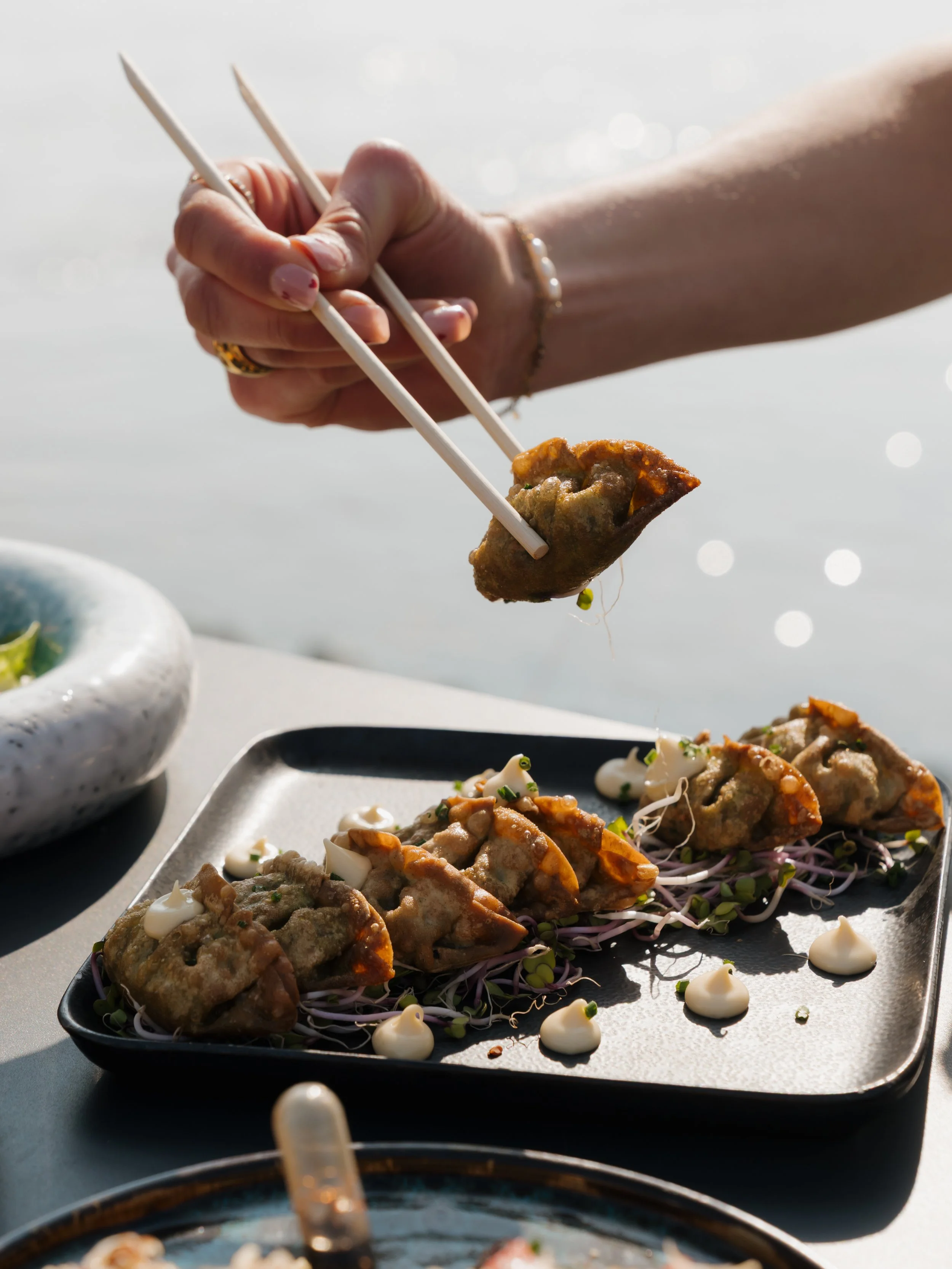 Close-up of a hand holding chopsticks picking up a piece of Asian dumpling or gyoza on a black rectangular plate, with more dumplings and small dollops of sauce on the plate.
