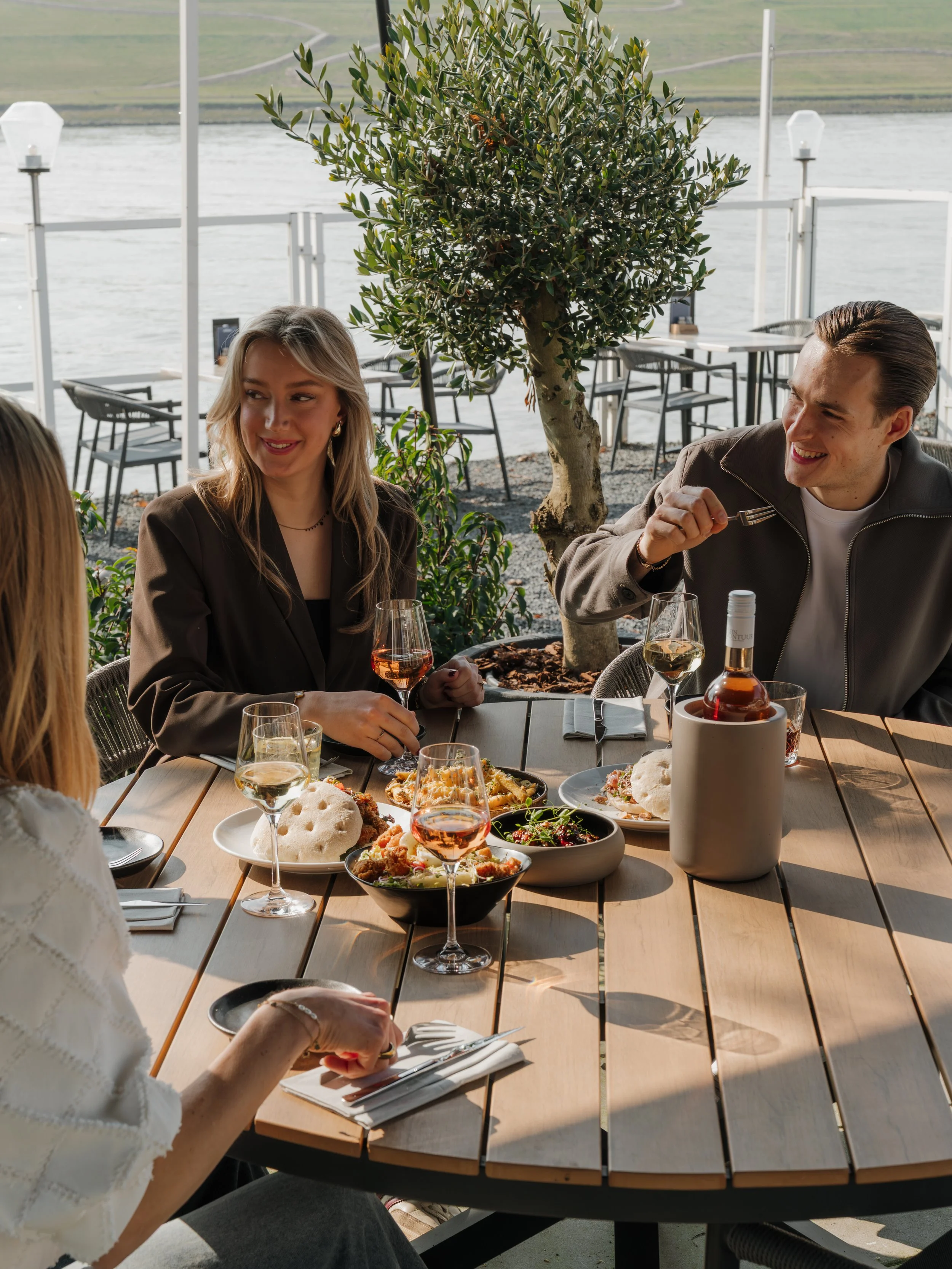 People enjoying a meal outdoors at a restaurant by the water, with plates of food and glasses of wine on the table.