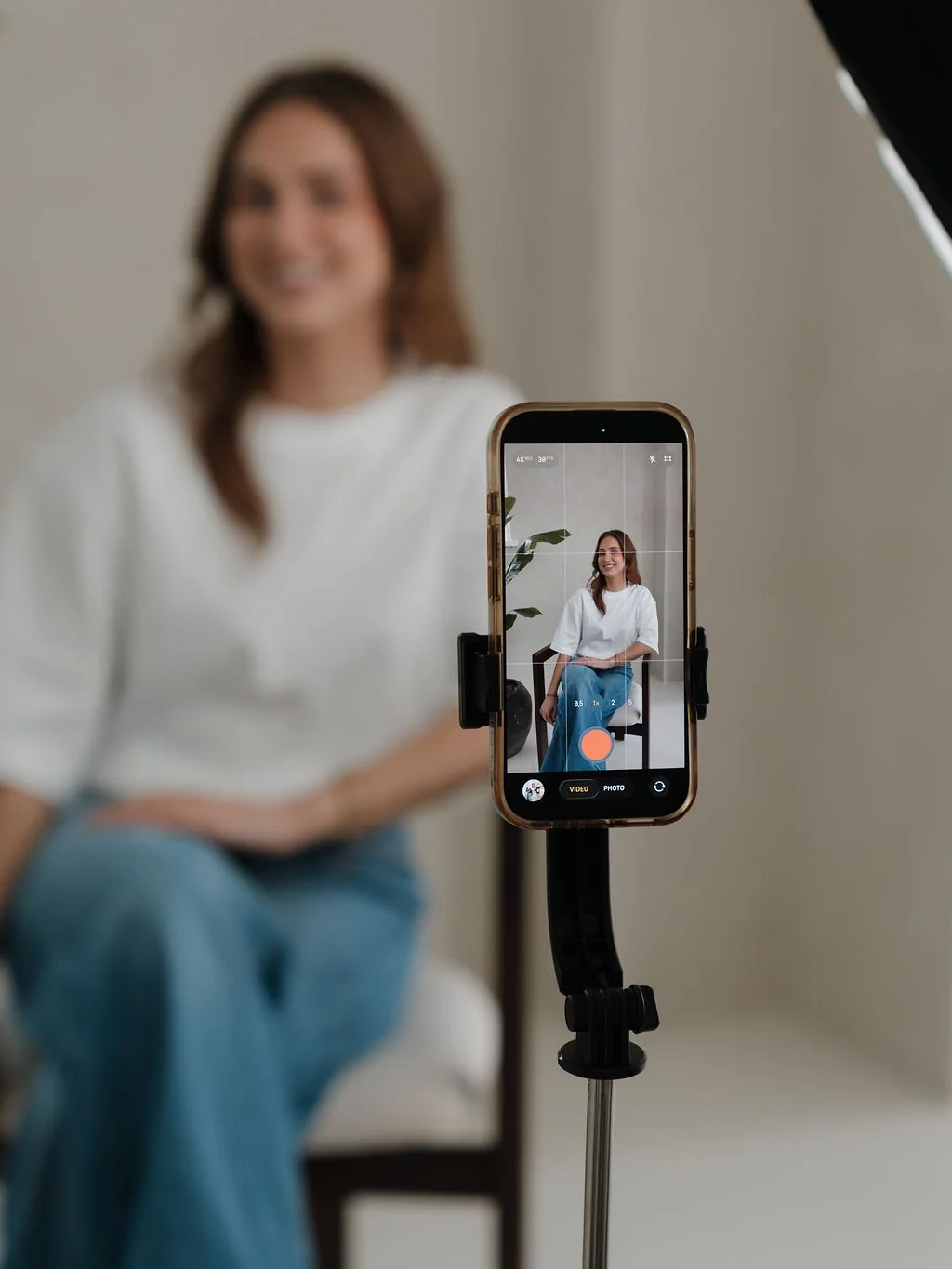 A woman is sitting on a chair in front of a camera on a tripod, taking a photo or video of herself with her smartphone.