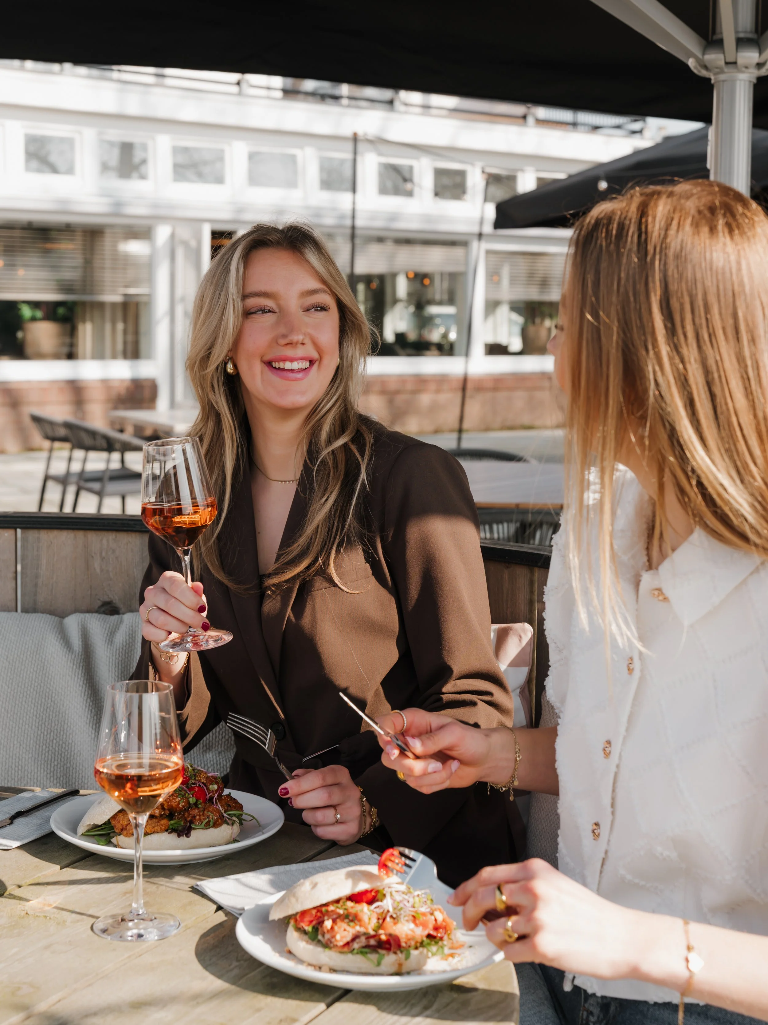 Two women enjoying a meal and drinks at an outdoor restaurant, one with blonde hair smiling and holding a glass of rosé, the other with reddish hair holding a fork and smiling.