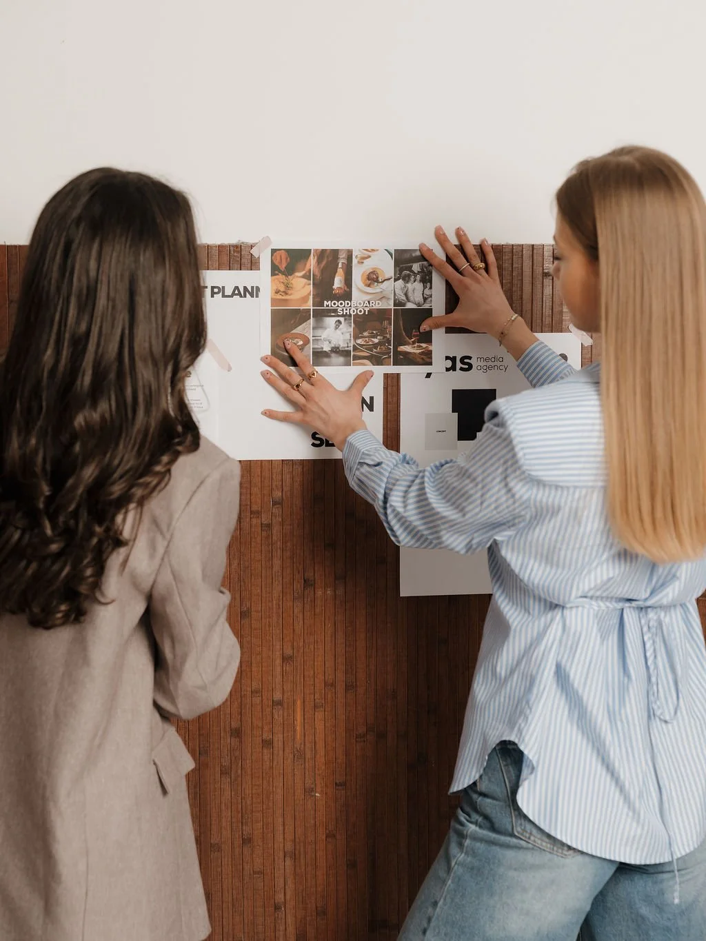 Two women discuss ideas and pin pictures on a corkboard for a media project or presentation.