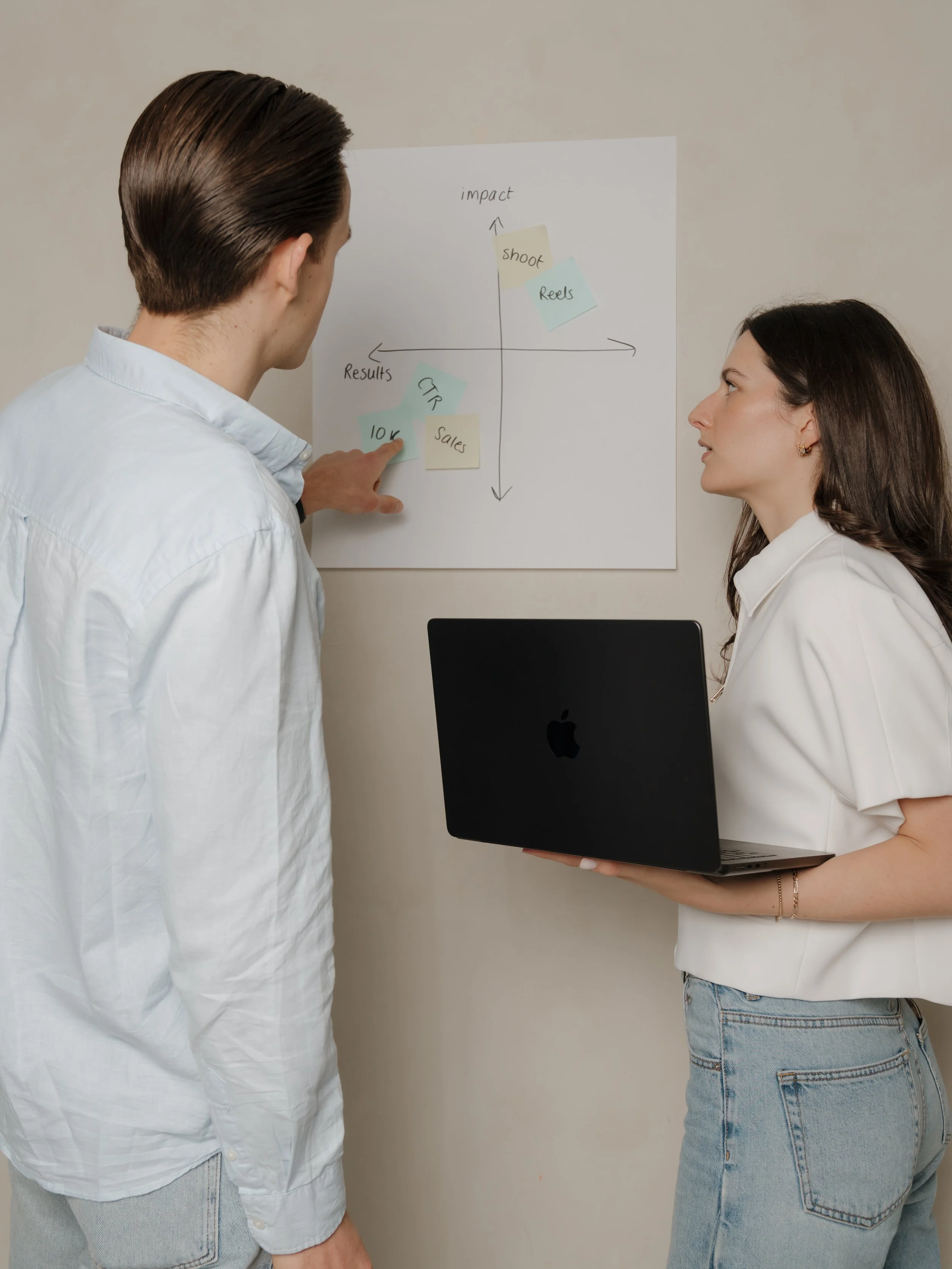 Two women are having a discussion in front of a whiteboard with various notes on it. One woman with brown hair, wearing a white shirt, is pointing at the notes. The other woman, with dark hair and holding a laptop, is listening. The notes on the whiteboard include words like "impact," "shoot," "reels," "results," "CTR," "10K," and "sales."