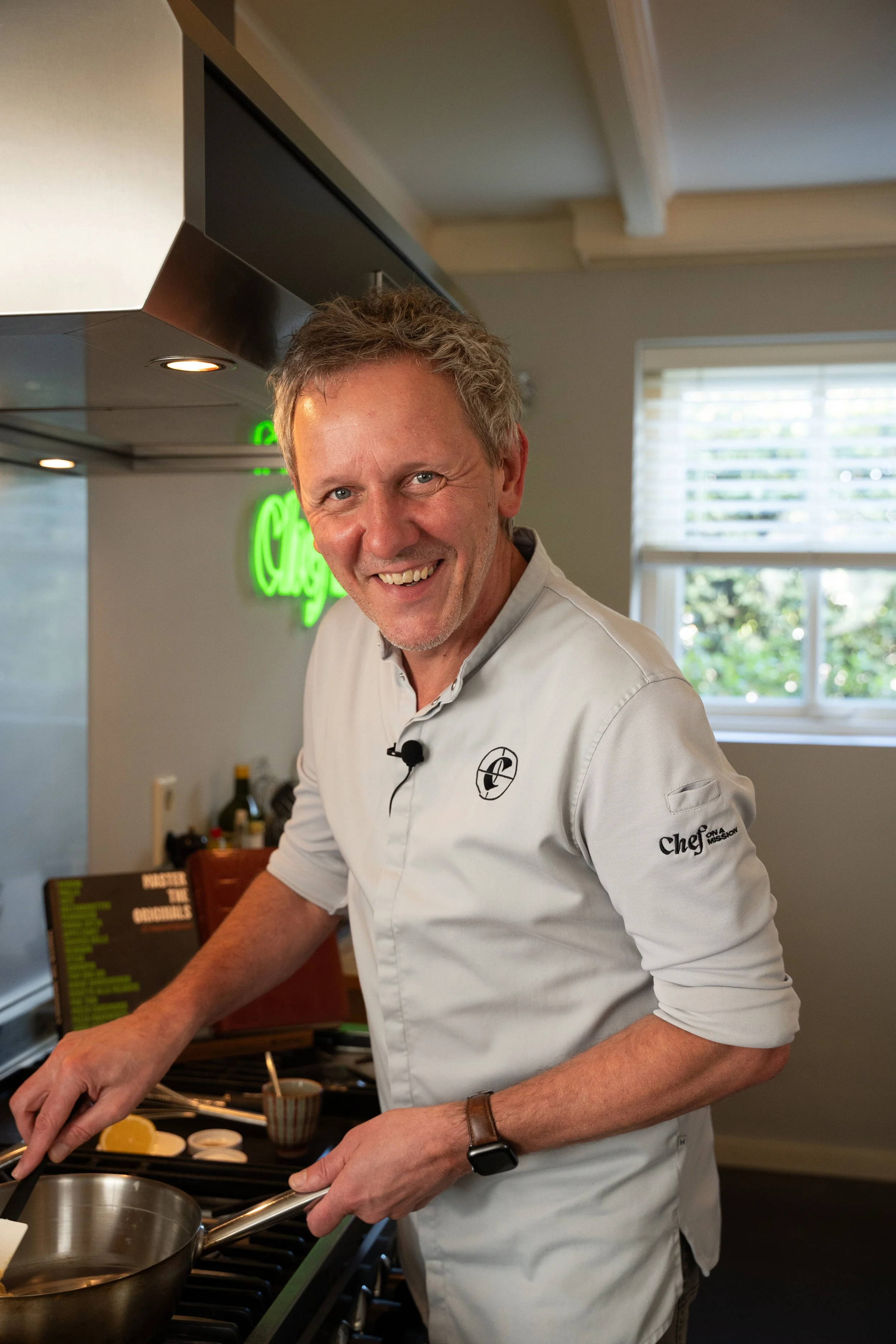 Male chef preparing food in a kitchen, smiling at the camera, wearing a white chef's coat, with a green illuminated sign in the background and a window showing trees outside.