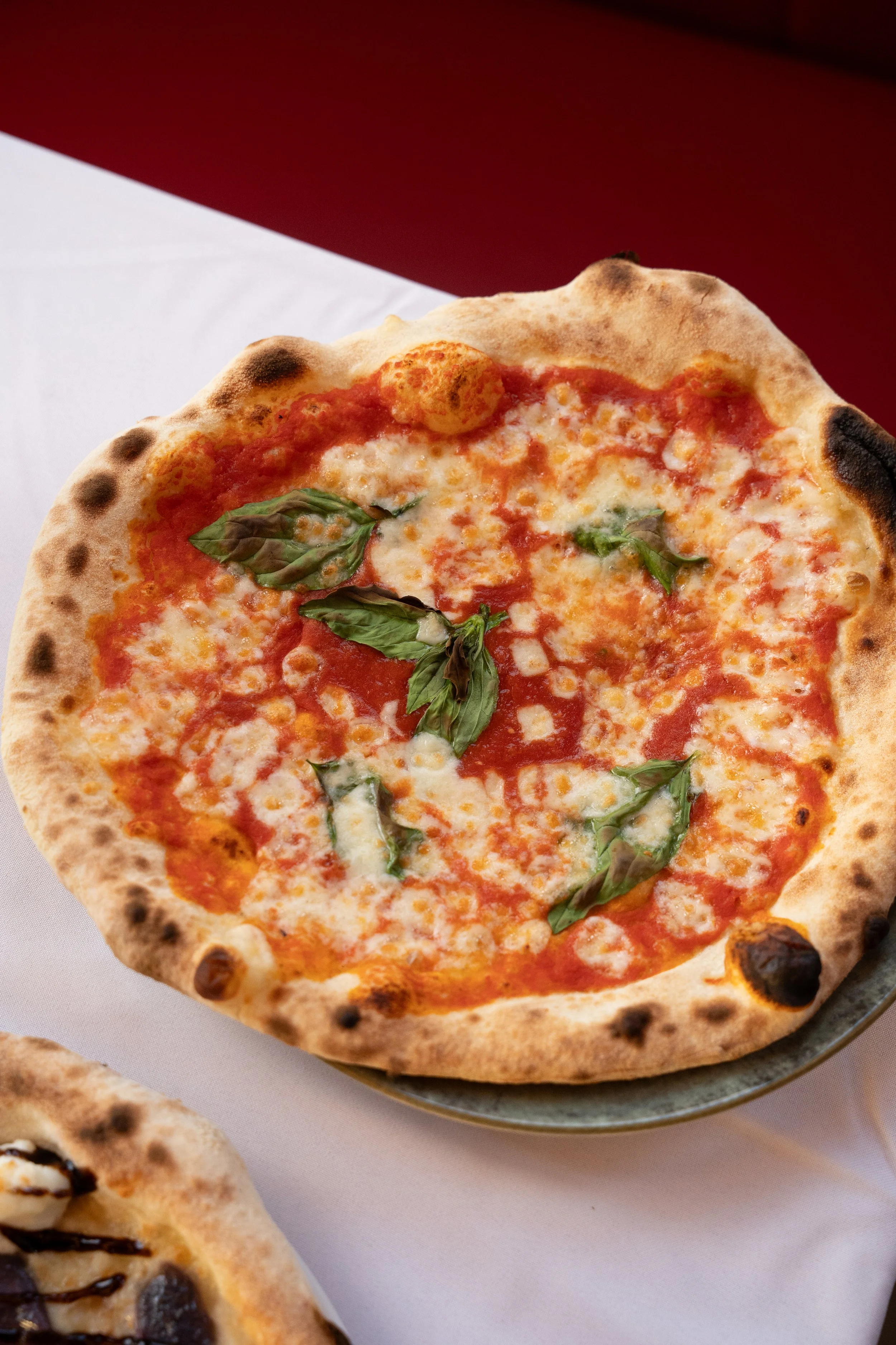 A close-up of a Margherita pizza with melted cheese, tomato sauce, and basil leaves on a plate, on a white tablecloth with a red background.