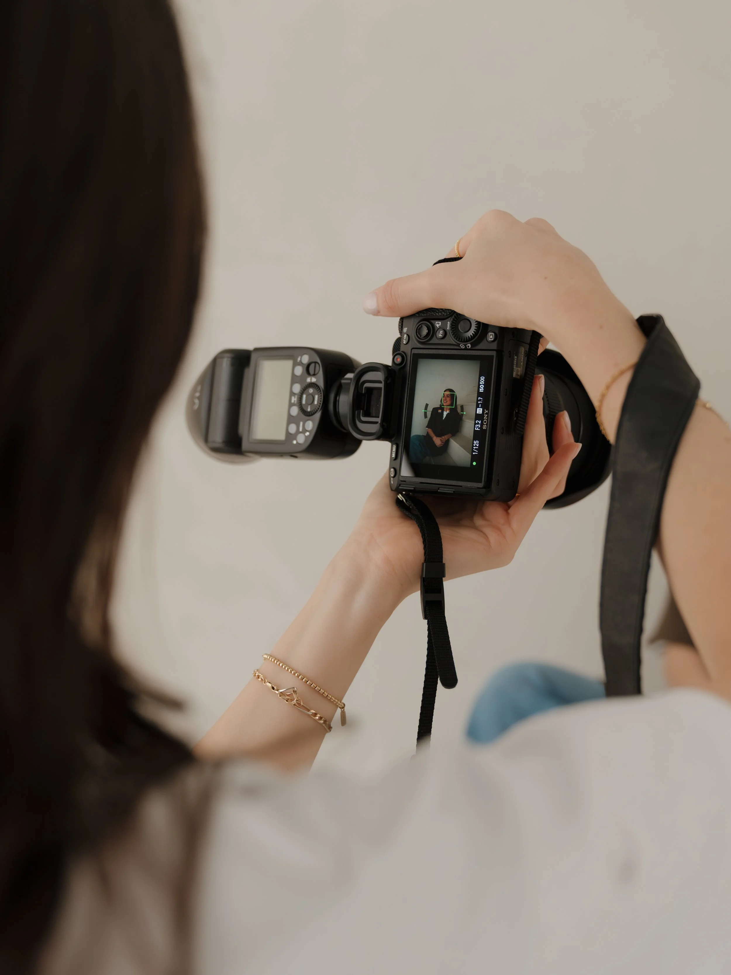 A woman taking a photo of herself with a DSLR camera, showing her reflection on the camera's LCD screen, against a plain background.